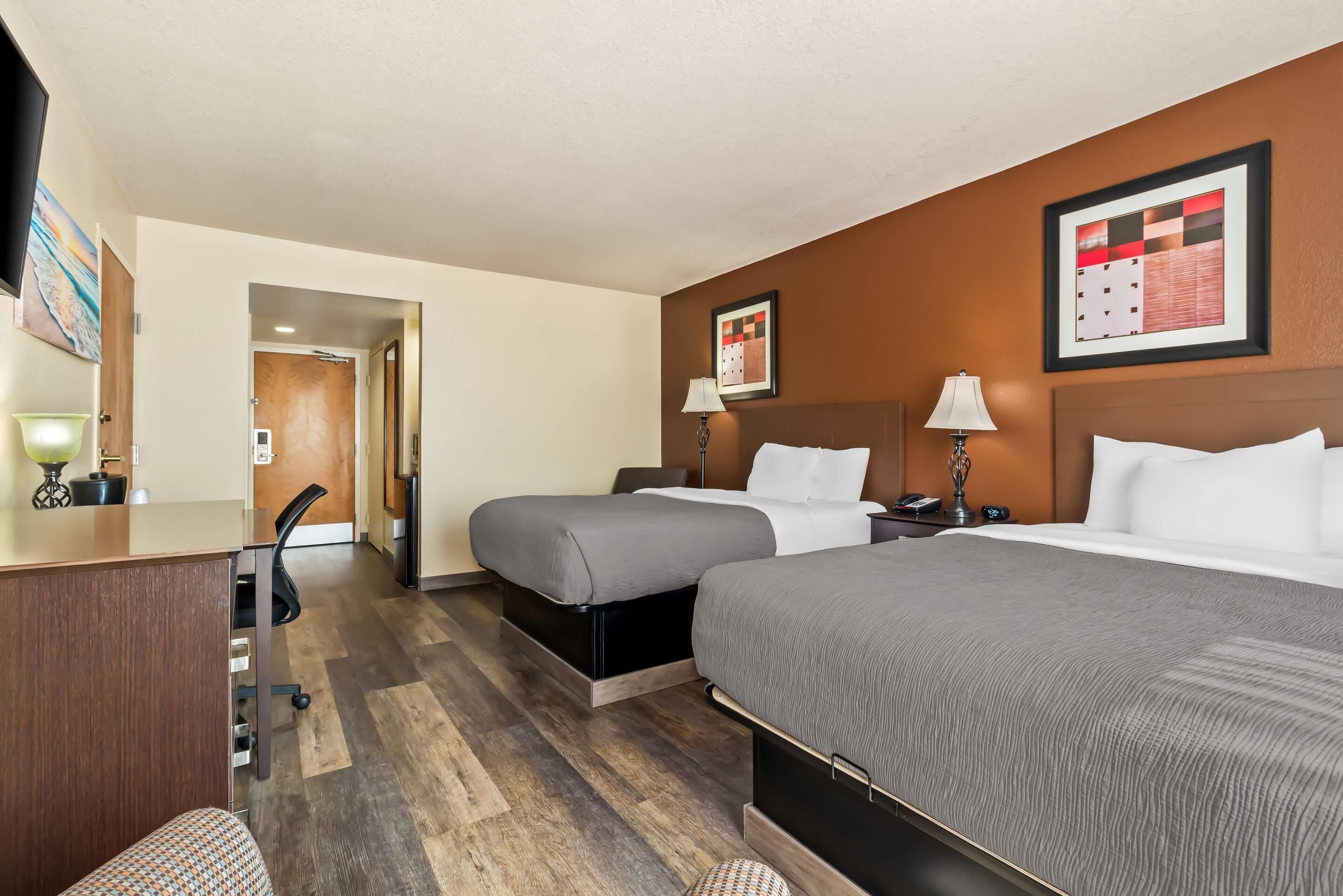 A hotel room with two beds, a desk, a chair, and a small table with a lamp.
