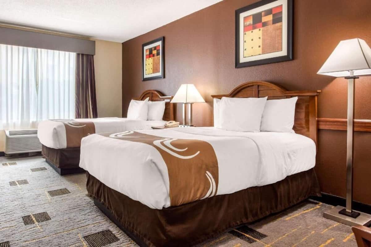 A hotel room with two beds, a large window with curtains, a standing lamp, and a framed artwork on the wall.