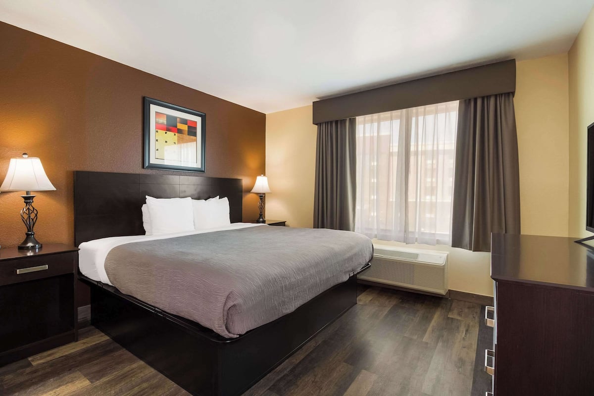 A hotel room with a large bed, two bedside tables with lamps, a framed artwork above the bed, a large window with curtains, and a built-in air conditioning unit.