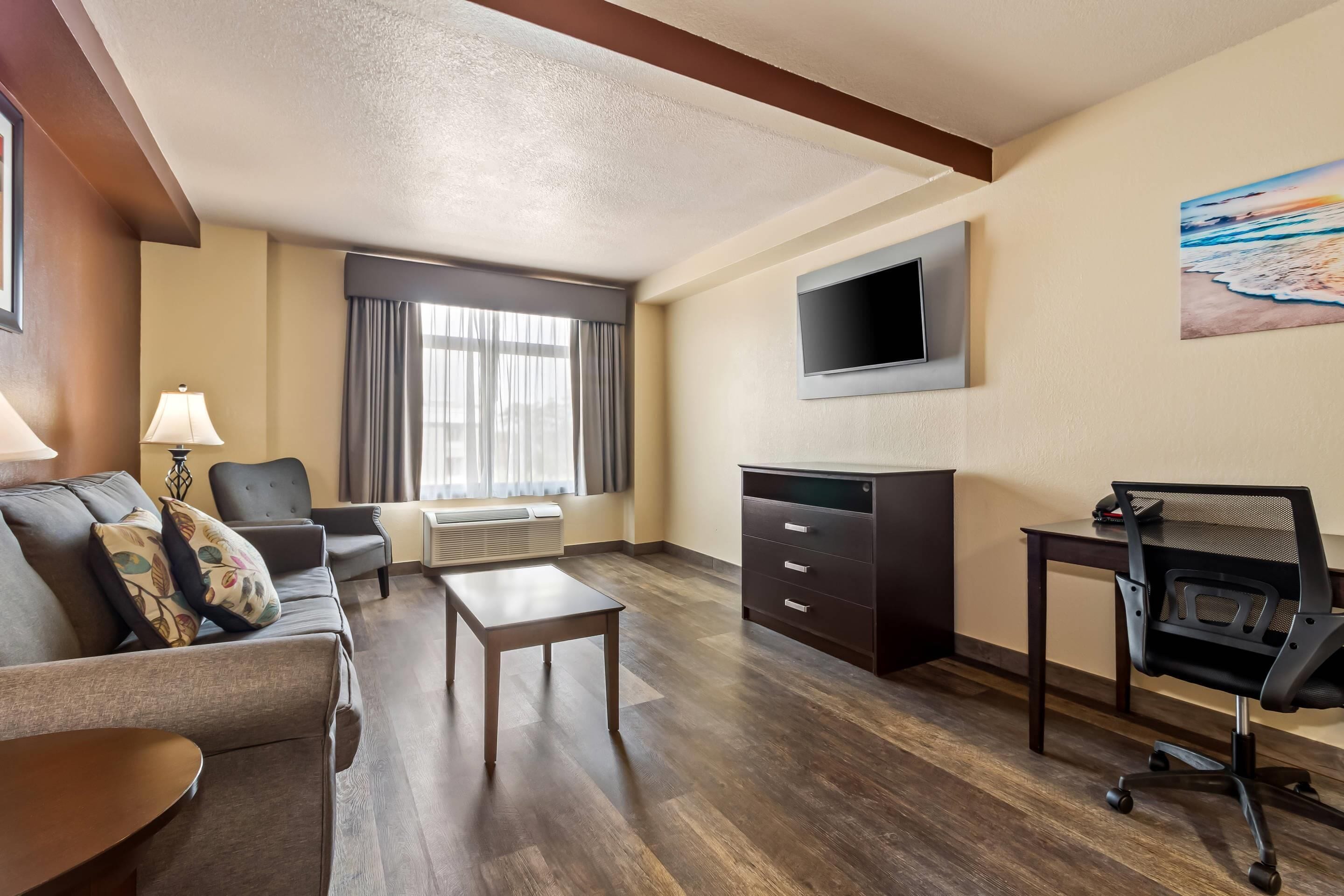A hotel room with a sofa, a small table, a desk with a chair, a TV mounted on the wall, and a window with curtains.