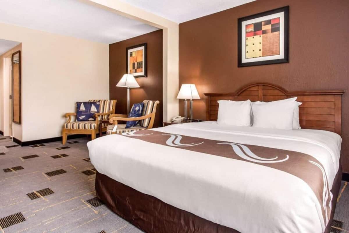 A hotel room with a large bed, two bedside lamps, a wooden headboard, a chair with a striped cushion, and a framed picture on the wall.