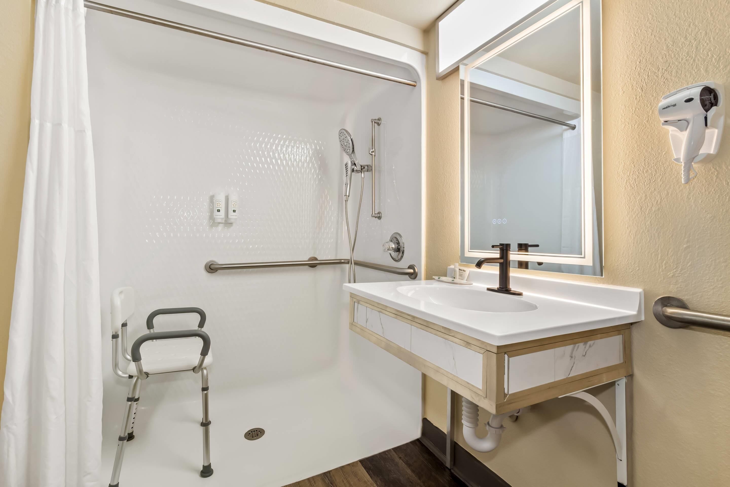 Bagno con doccia, lavandino, specchio, asciugatore a mano e sedia a rotelle.