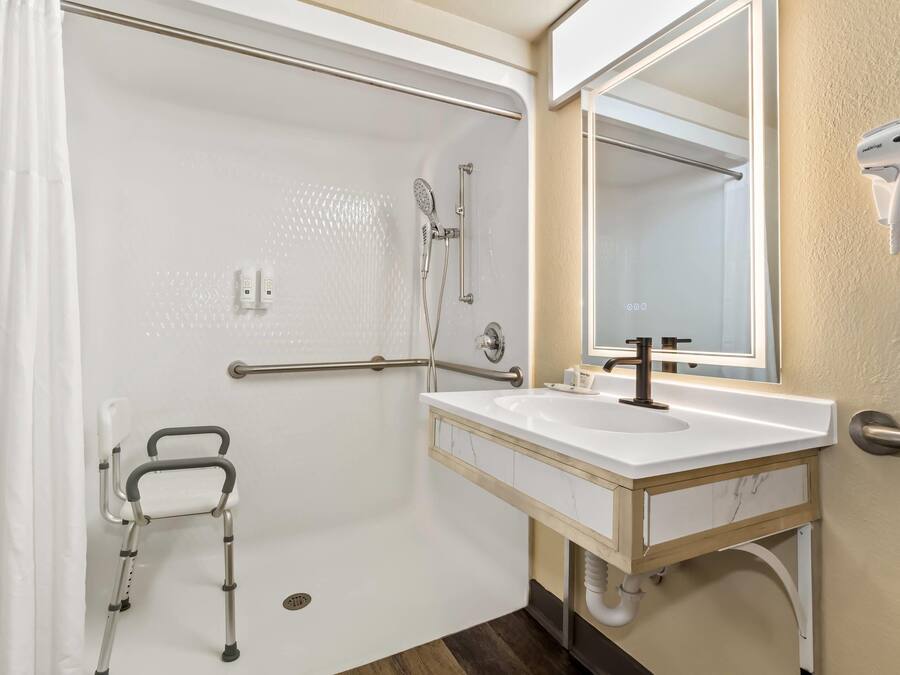 Bagno con doccia, lavandino, specchio, asciugatore a mano e sedia a rotelle.