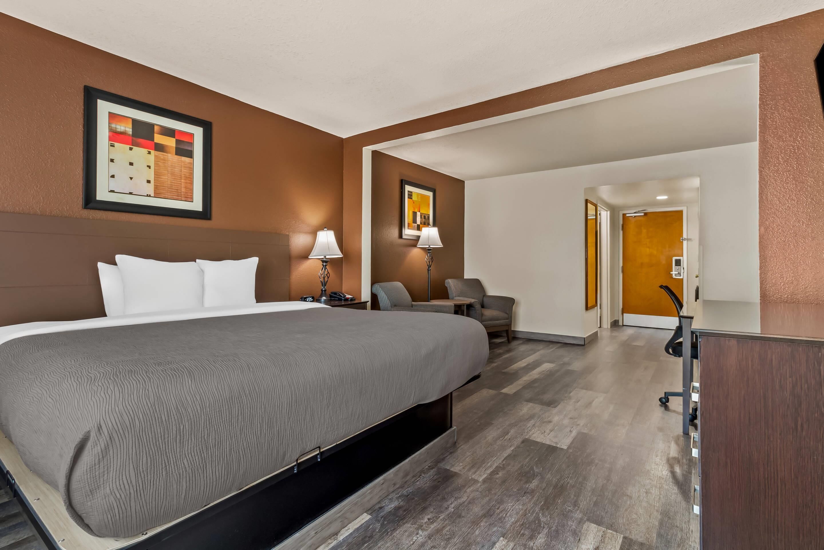 A hotel room with a large bed, two bedside tables with lamps, a desk, and a chair. There is a door leading to another room.