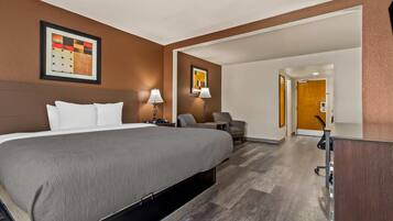 A hotel room with a large bed, two bedside tables with lamps, a desk, and a chair. There is a door leading to another room.
