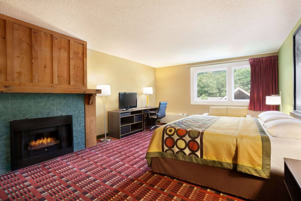 A hotel room with a fireplace, a bed with a patterned bedspread, a desk with a computer, and a window with curtains.