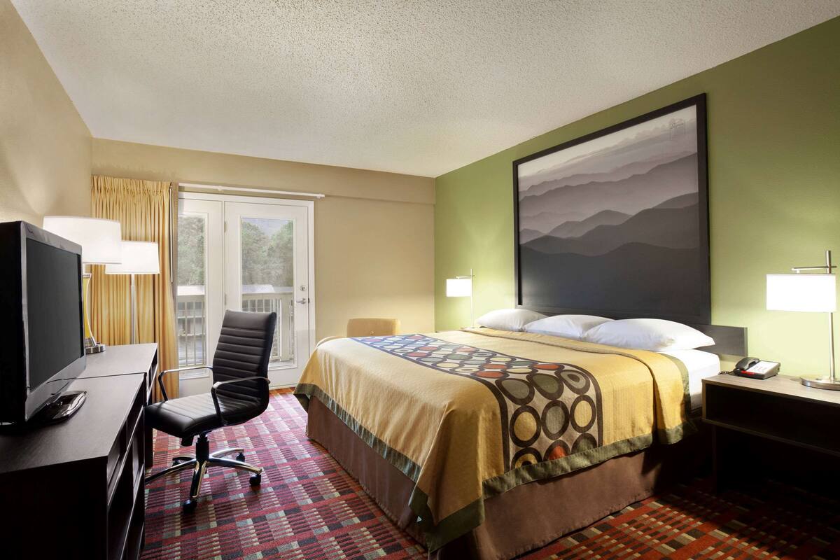 A hotel room with a bed, a desk with a computer, a chair, a TV, and a window with curtains.