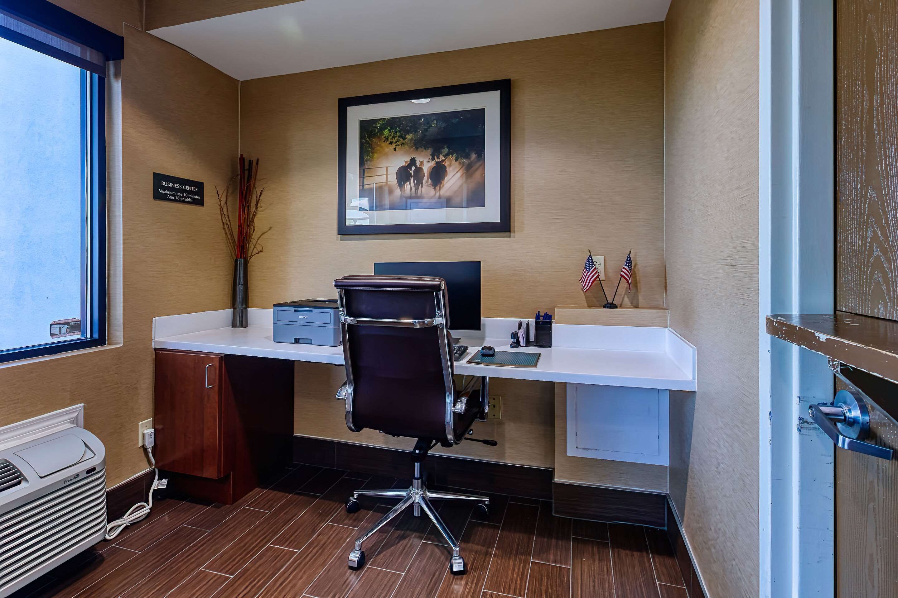 A business center with a desk, computer, and office chair. There is a framed picture on the wall, an American flag, and a sign indicating the business center location.