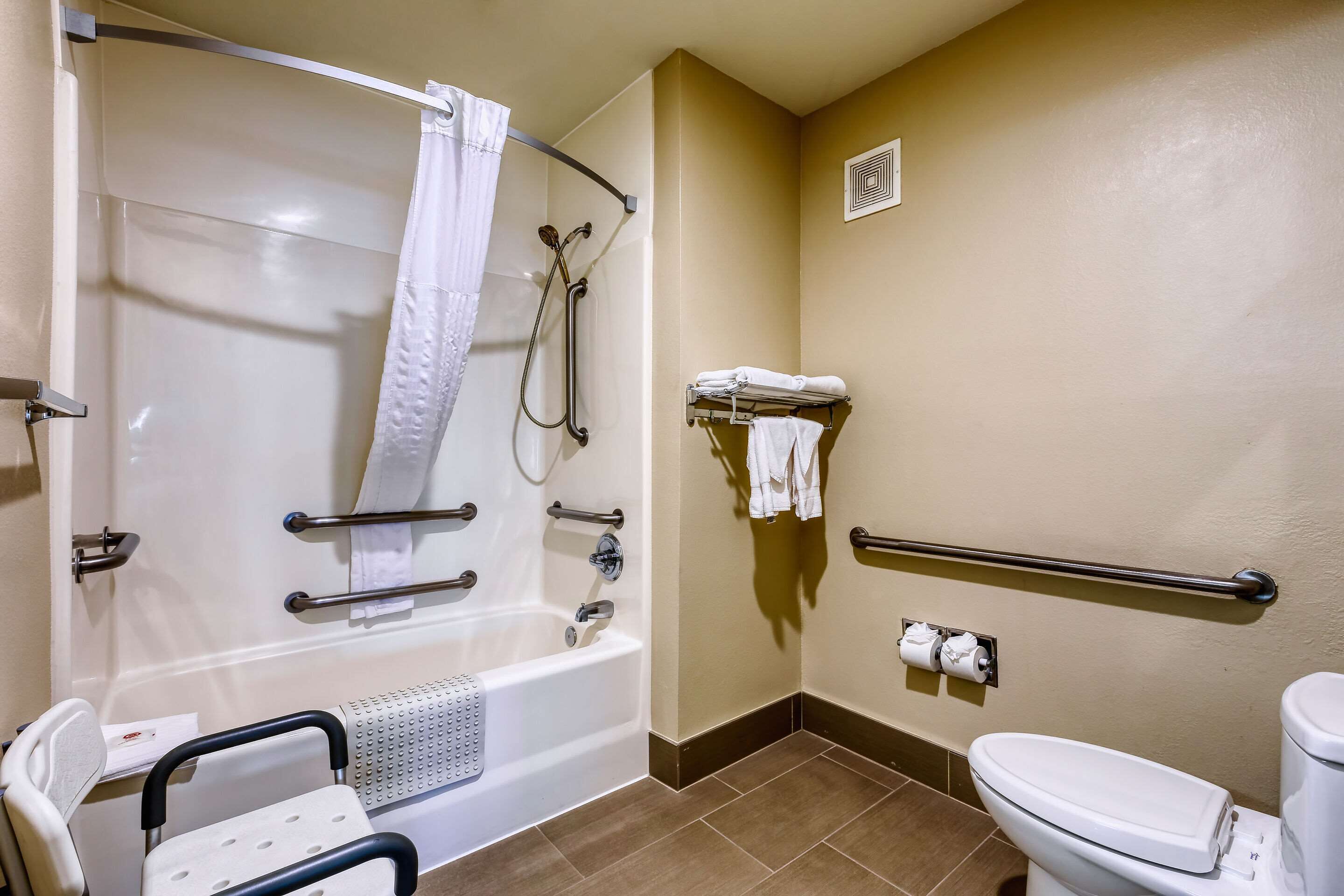 A bathroom with a bathtub, a toilet, a shower with a grab bar, a wheelchair-accessible chair, and a towel rack.
