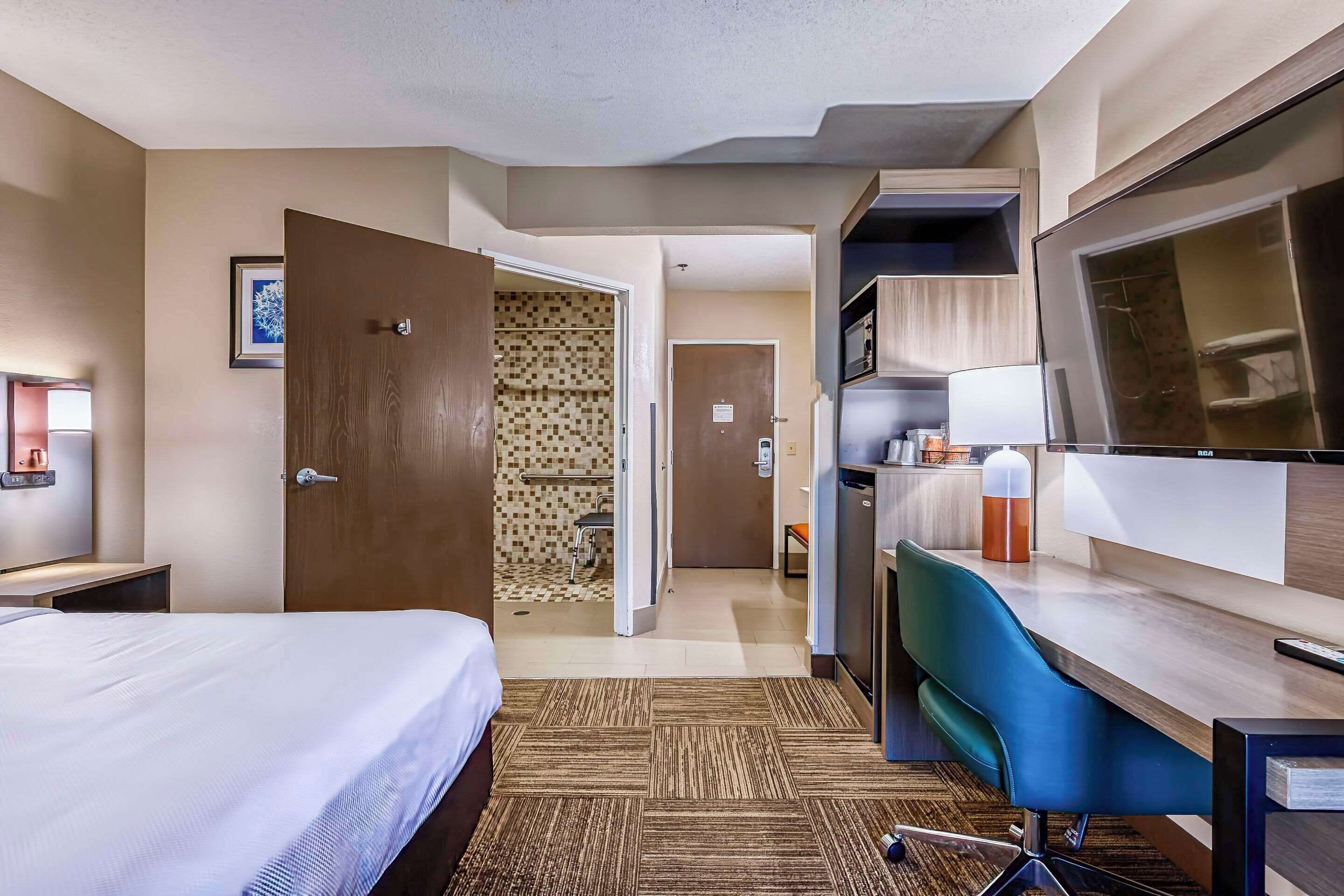 A hotel room with a bed, a desk with a chair, a television, and a bathroom visible through an open door.