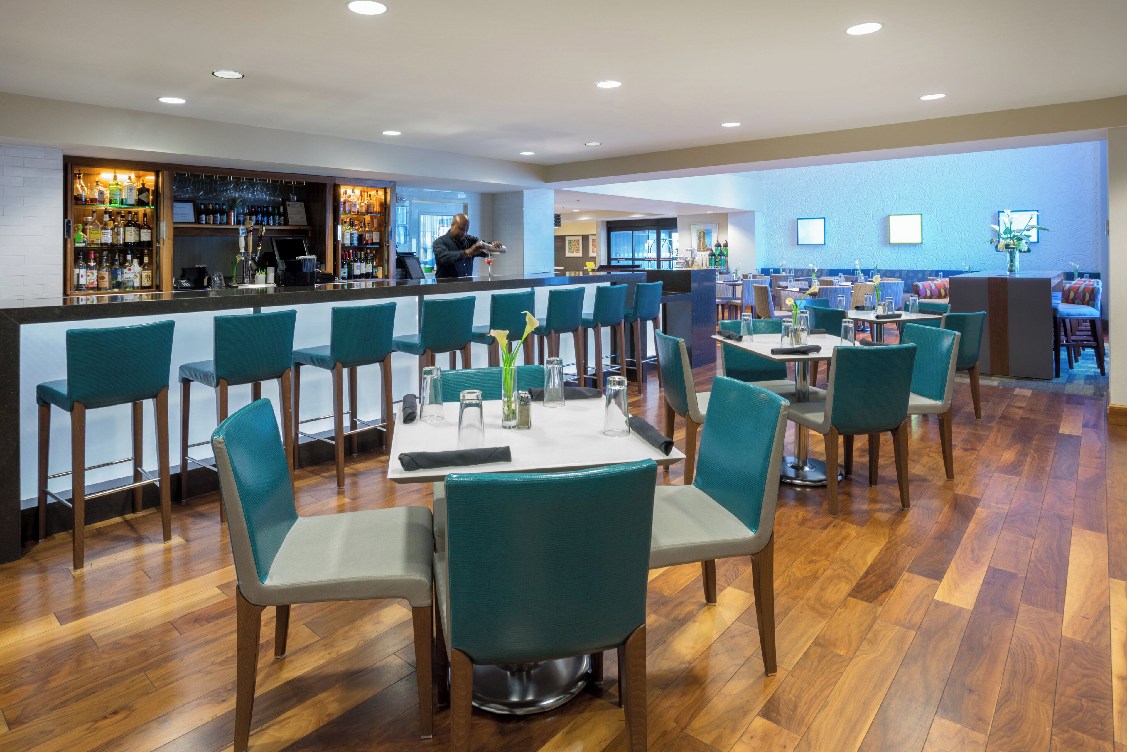 Un'area bar con un barista, una zona salotto con tavoli e sedie, e una parete decorata.