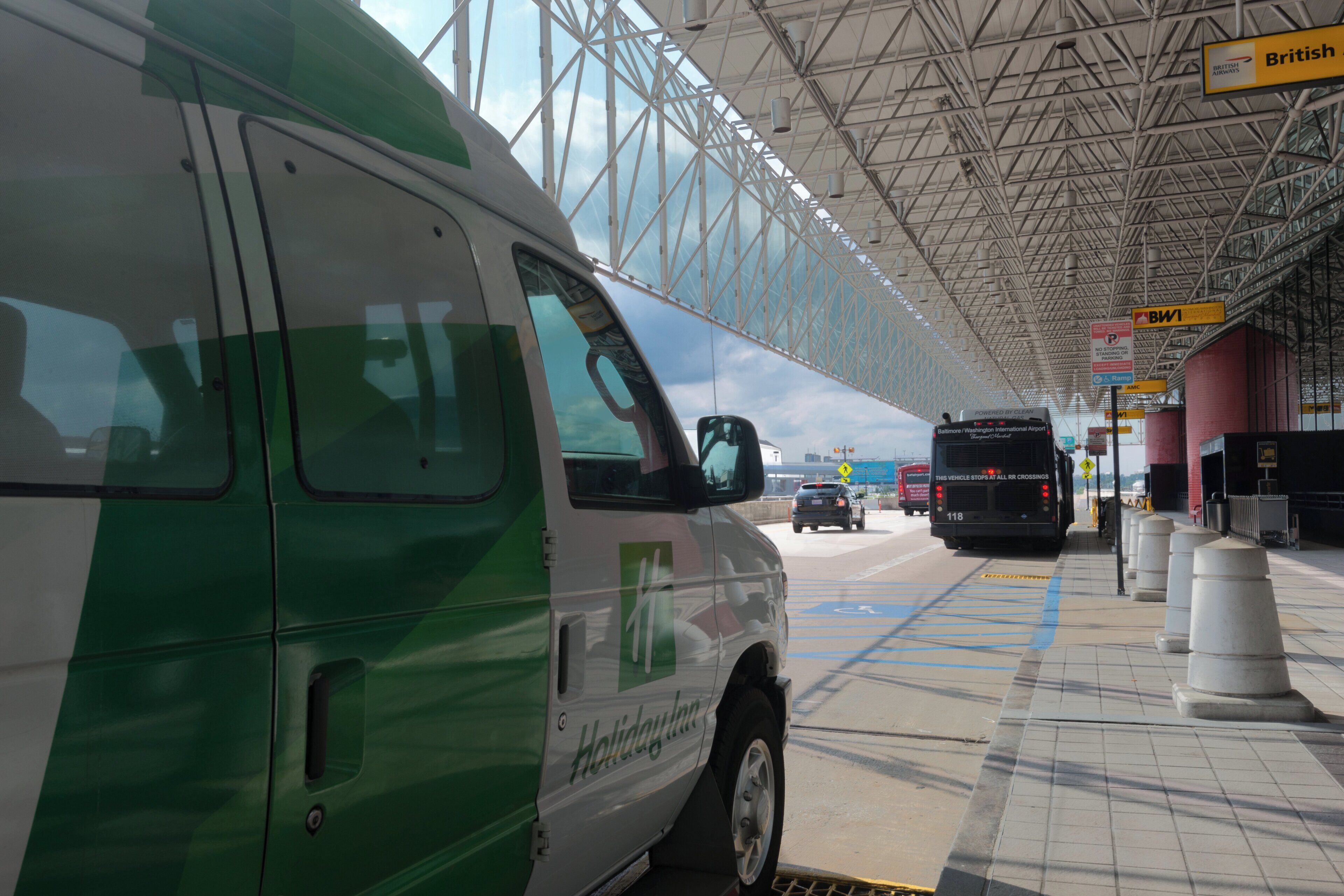 A green and white van parked at a bus station with a clear sky and a metal roof structure.