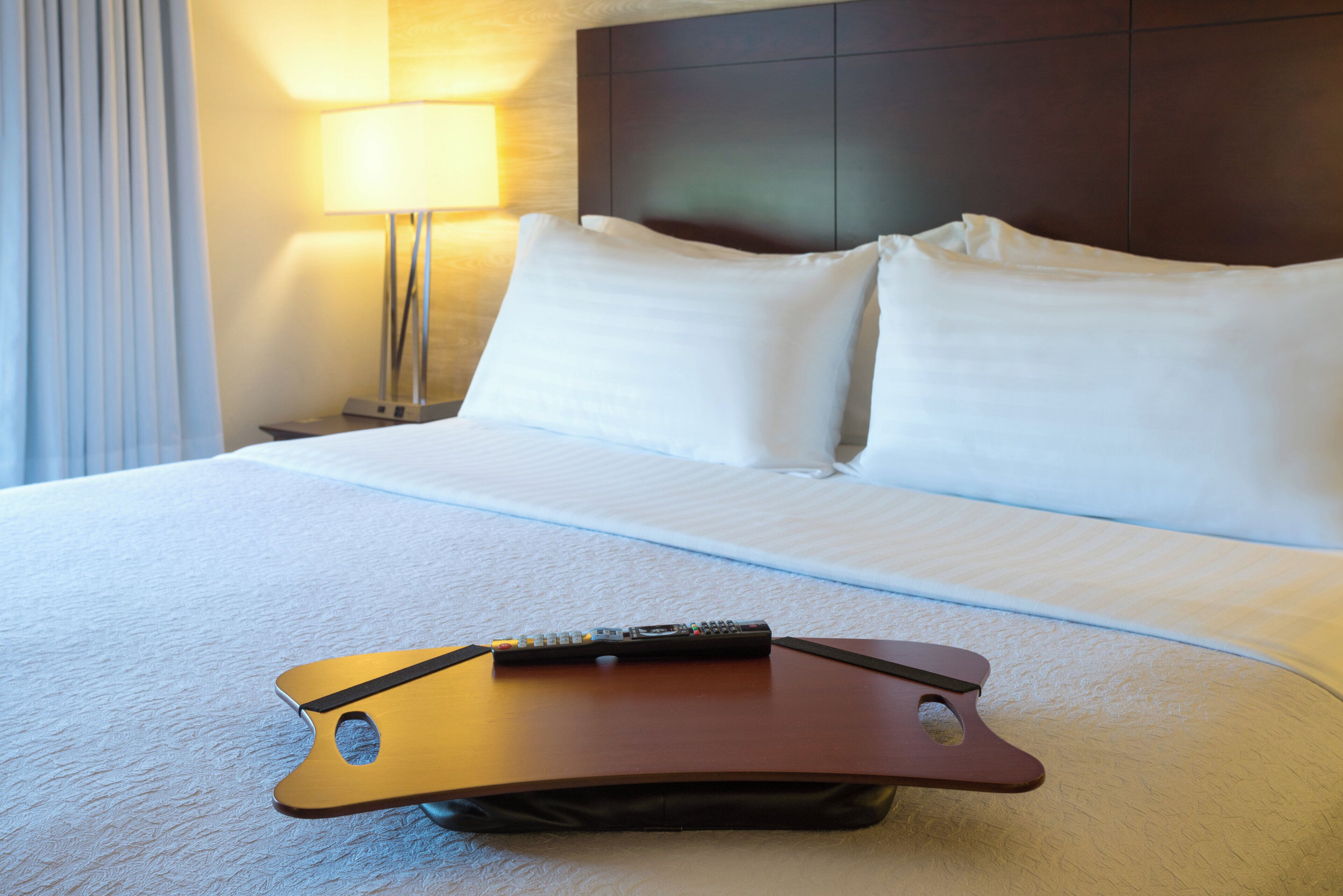 A hotel room with a bed, pillows, a remote control, and a lamp.