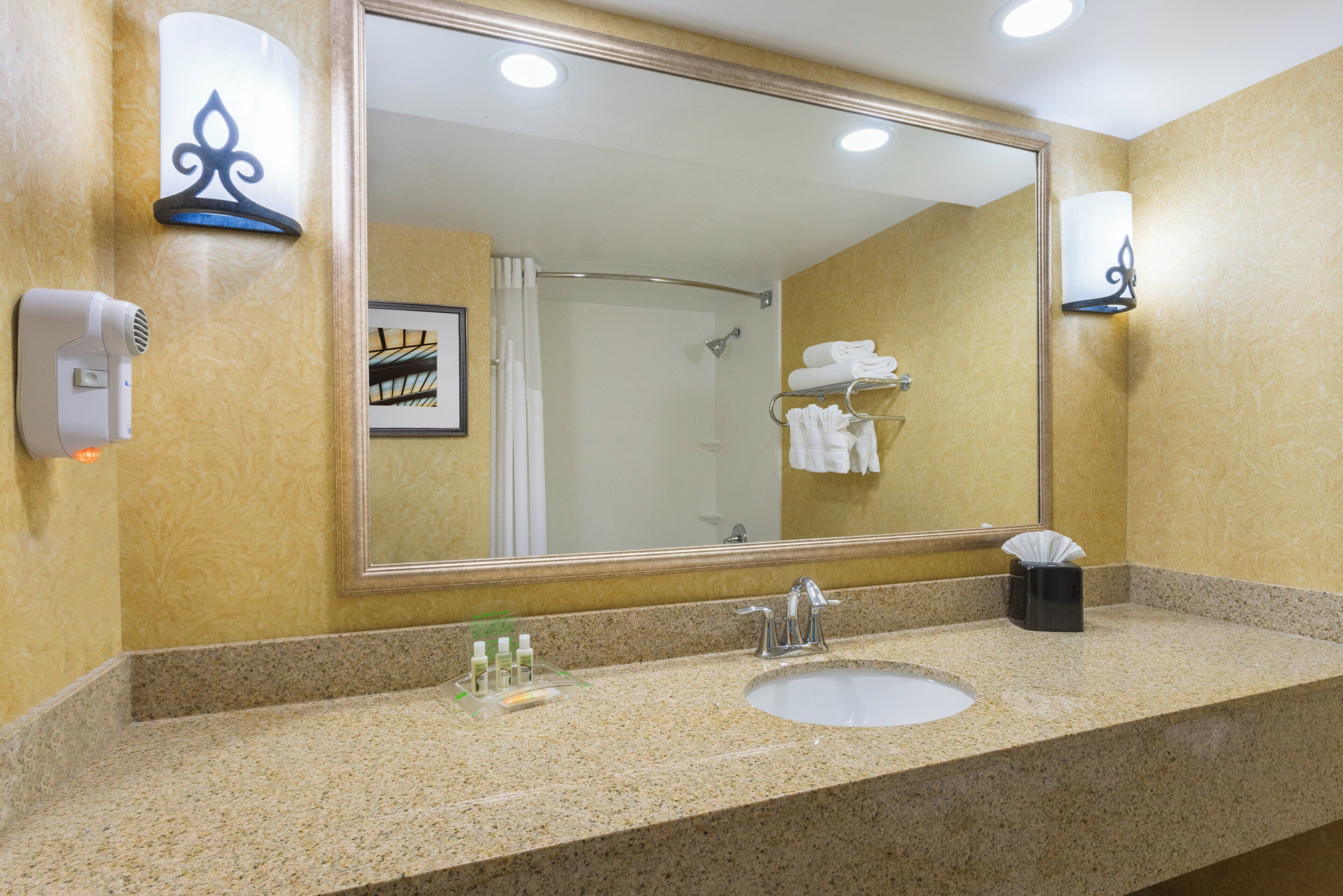 A hotel bathroom with a granite countertop, a sink, a mirror, a towel rack, and a hand dryer.