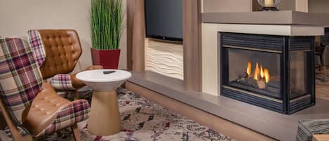 A modern living room with a fireplace, a television mounted on the wall, a plaid chair, a small round table, and a potted plant.