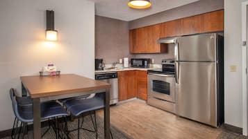 A compact kitchen with wooden cabinets, a stainless steel refrigerator, a microwave, and a dishwasher. There is a wooden table with four chairs and a small rug on the floor.