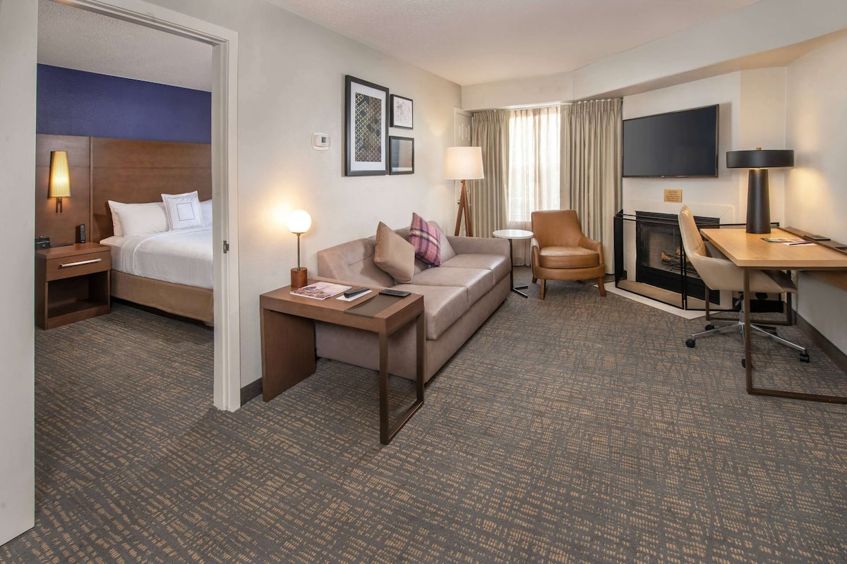 A hotel room with a bed, a sofa, a desk, a chair, a TV, and a fireplace.