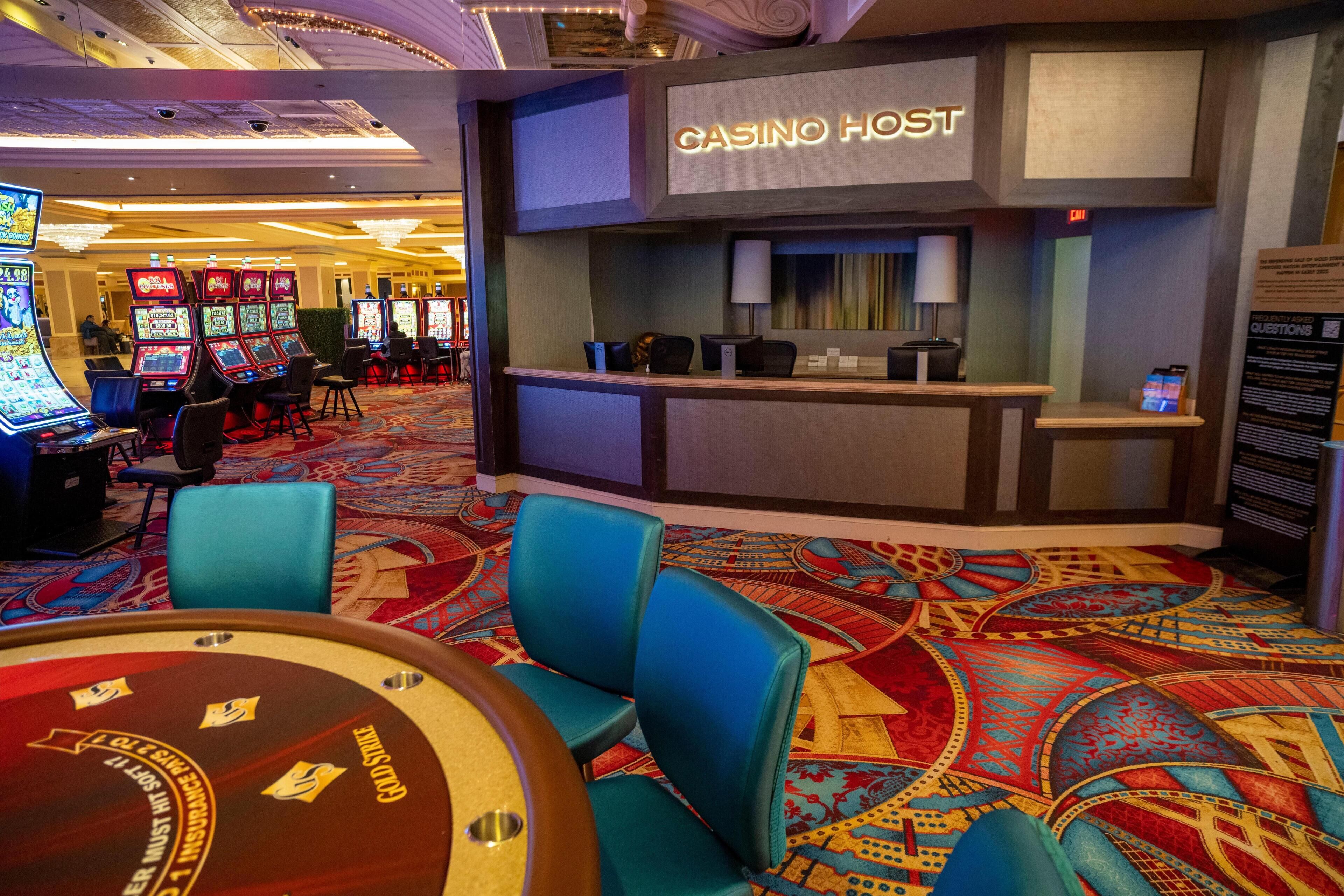 Ein Casino-Bereich mit Spielautomaten, einem Roulette-Tisch und einem Empfangsschalter mit der Aufschrift 'CASINO HOST'.