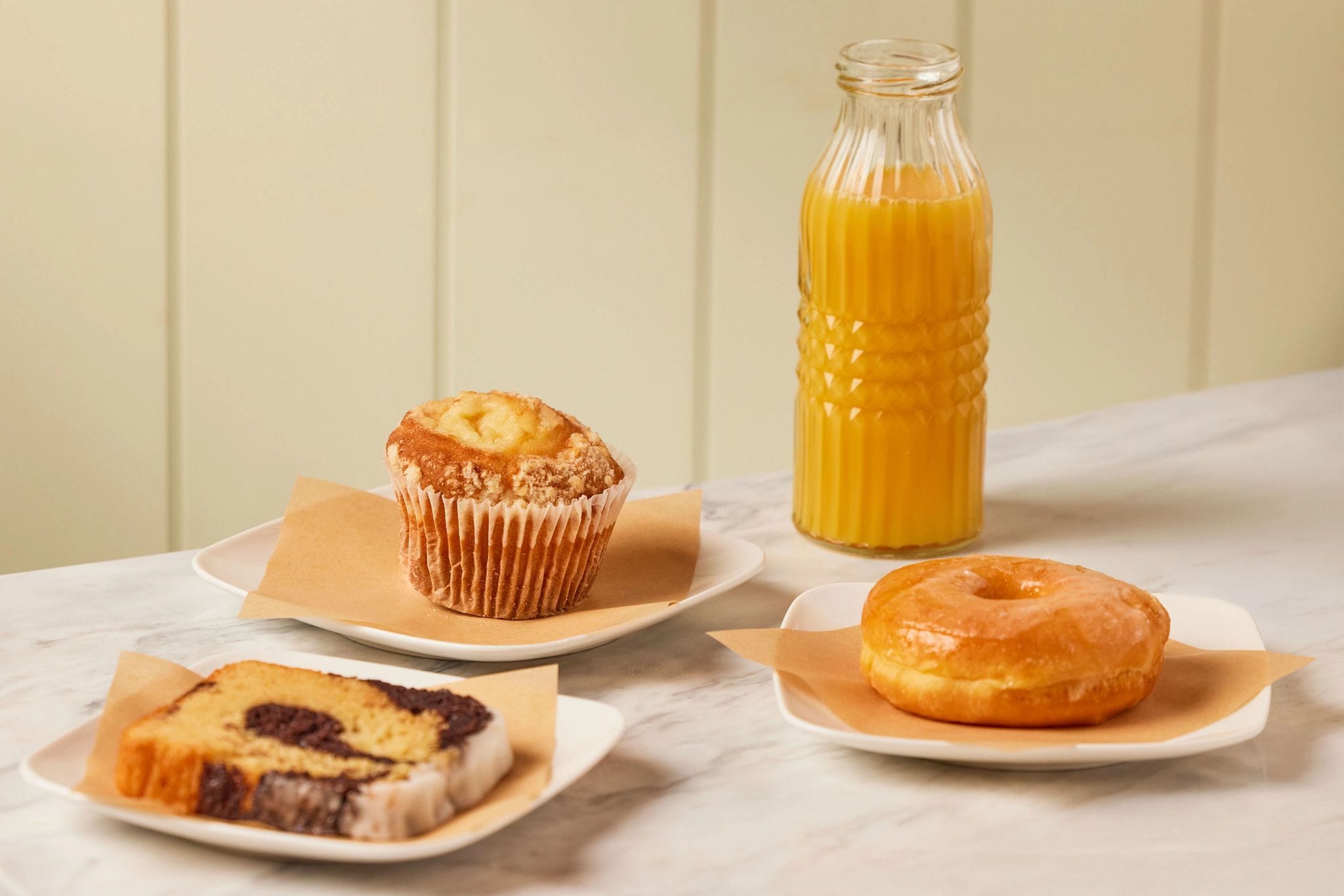 A marble countertop with a muffin, a donut, and a slice of cake on separate plates, and a glass bottle of orange juice.