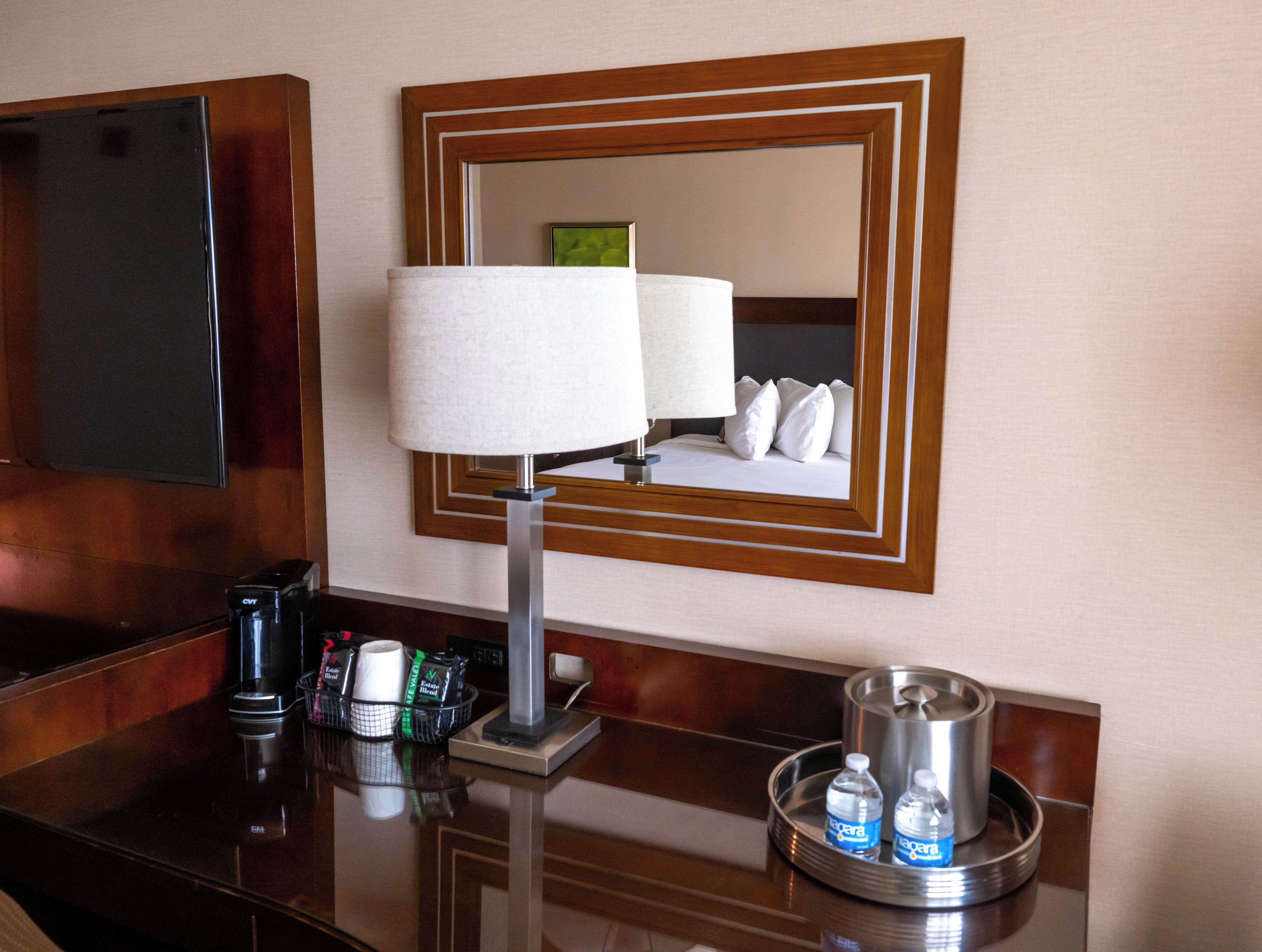 Habitación de hotel con mesita de noche, lámpara, espejo y bandeja con botellas de agua.
