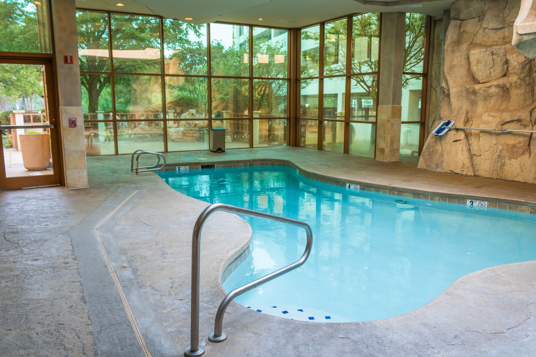 An indoor swimming pool with clear glass walls, a stone wall, and a metal pool ladder.