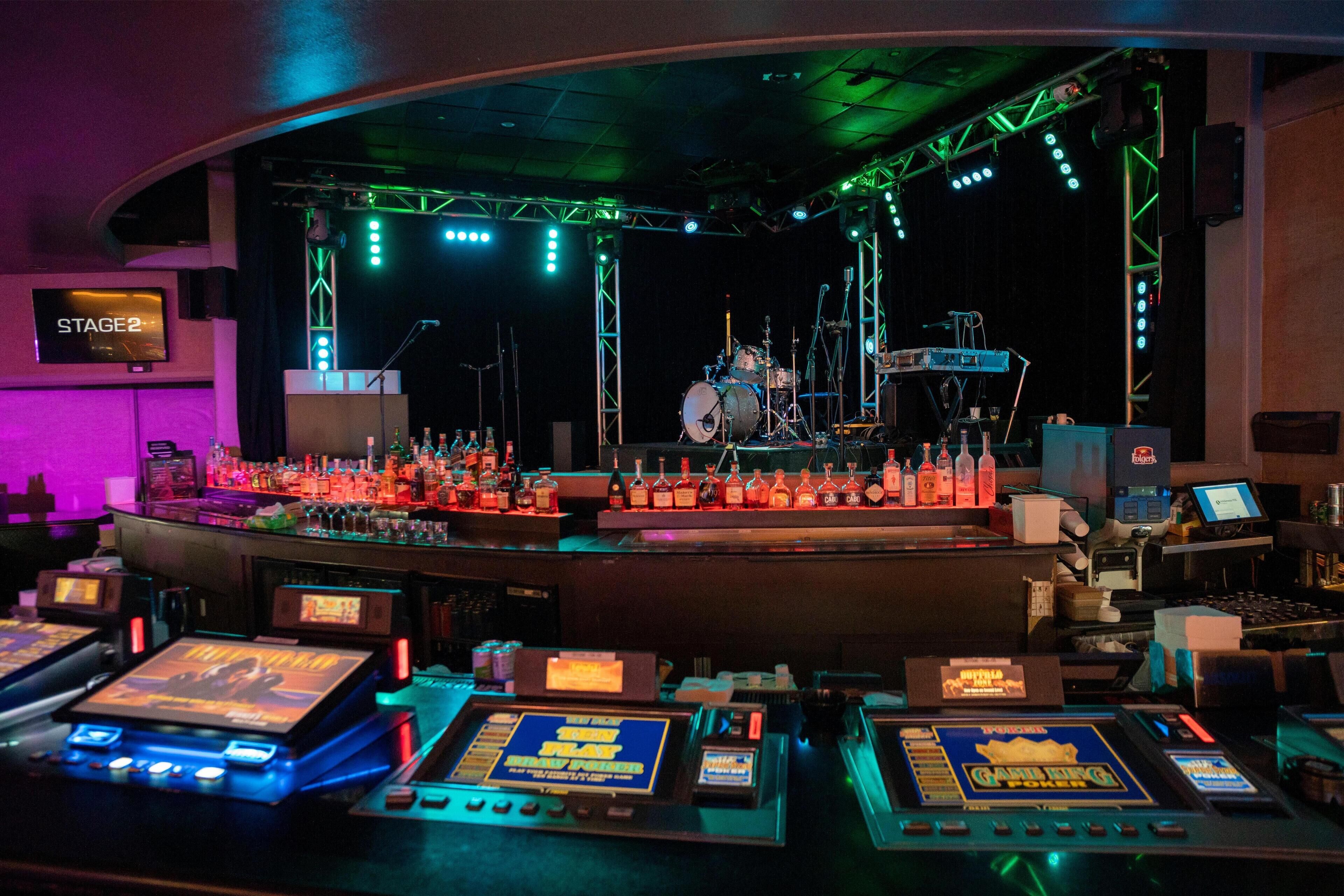 A stage with a drum set and lighting equipment, a bar area with bottles and gaming machines, and a sign reading "STAGE 2".