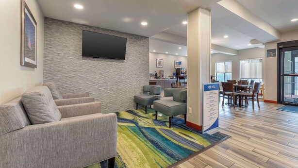 A hotel lobby with a seating area, a TV mounted on the wall, a menu stand, and a dining area with tables and chairs.
