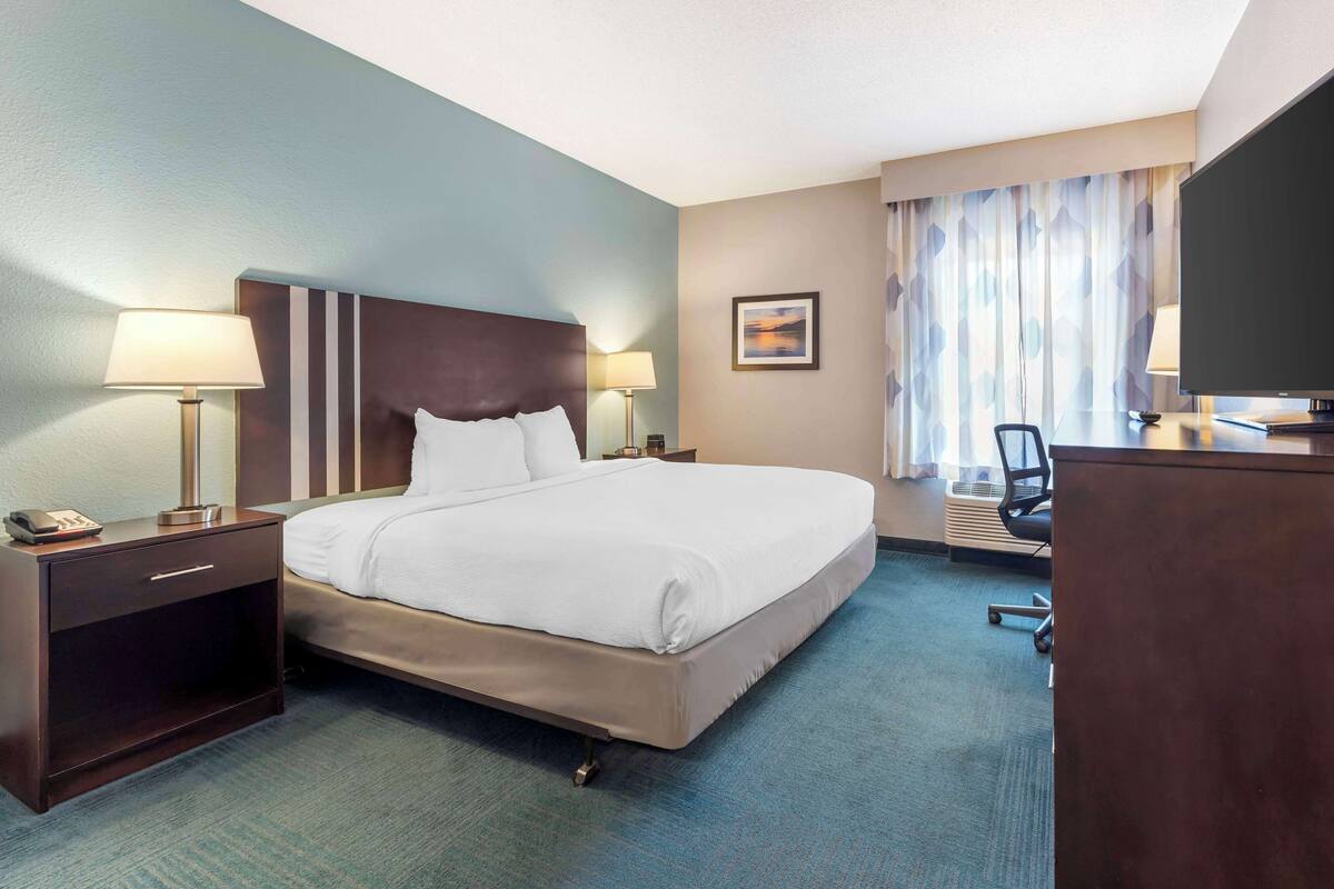Una habitación de hotel con una cama grande, dos mesitas de noche con lámparas, un escritorio con silla, un televisor y una ventana con cortinas.
