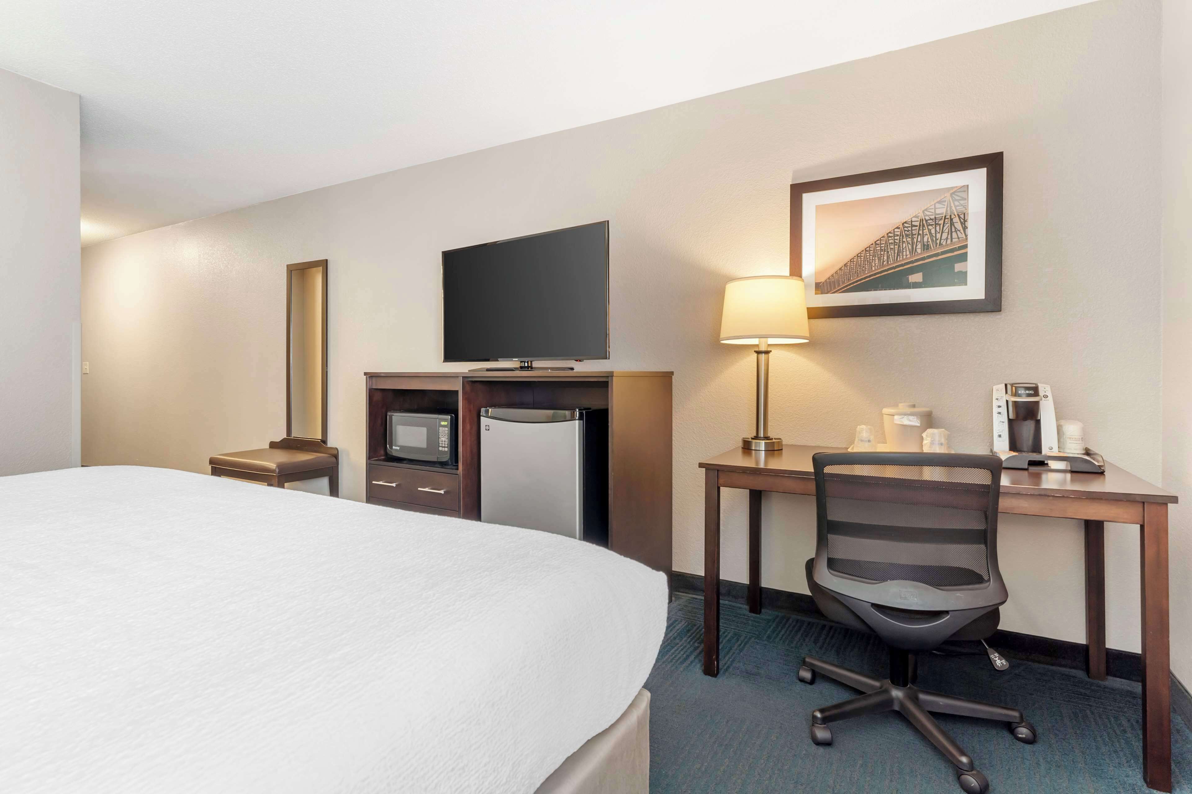 A hotel room with a bed, a desk with a computer, a chair, a television, a refrigerator, a microwave, a coffee maker, and a framed picture on the wall.