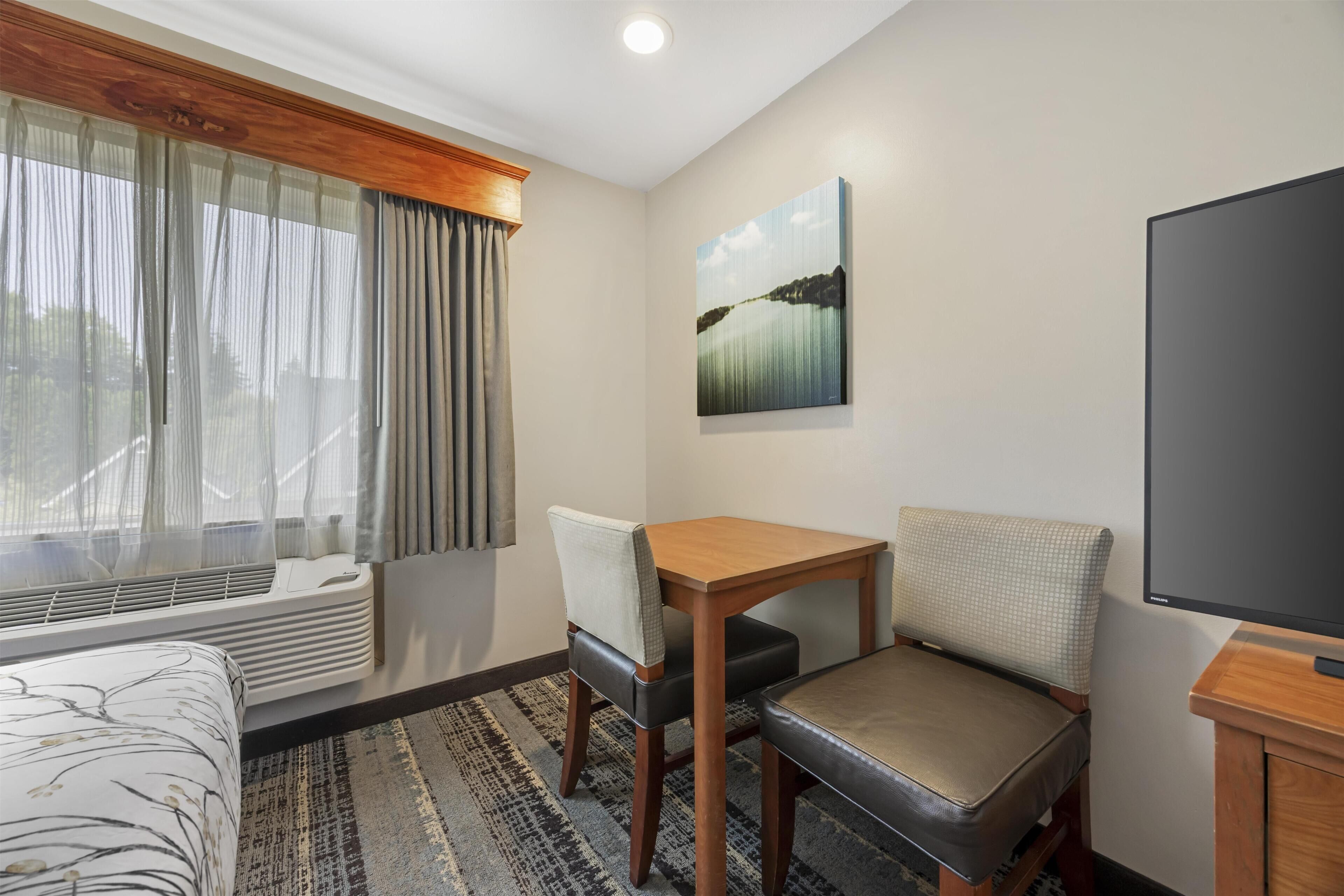 Habitación de hotel con cama, una mesa, dos sillas, televisión, un cuadro en la pared y una ventana con cortinas.