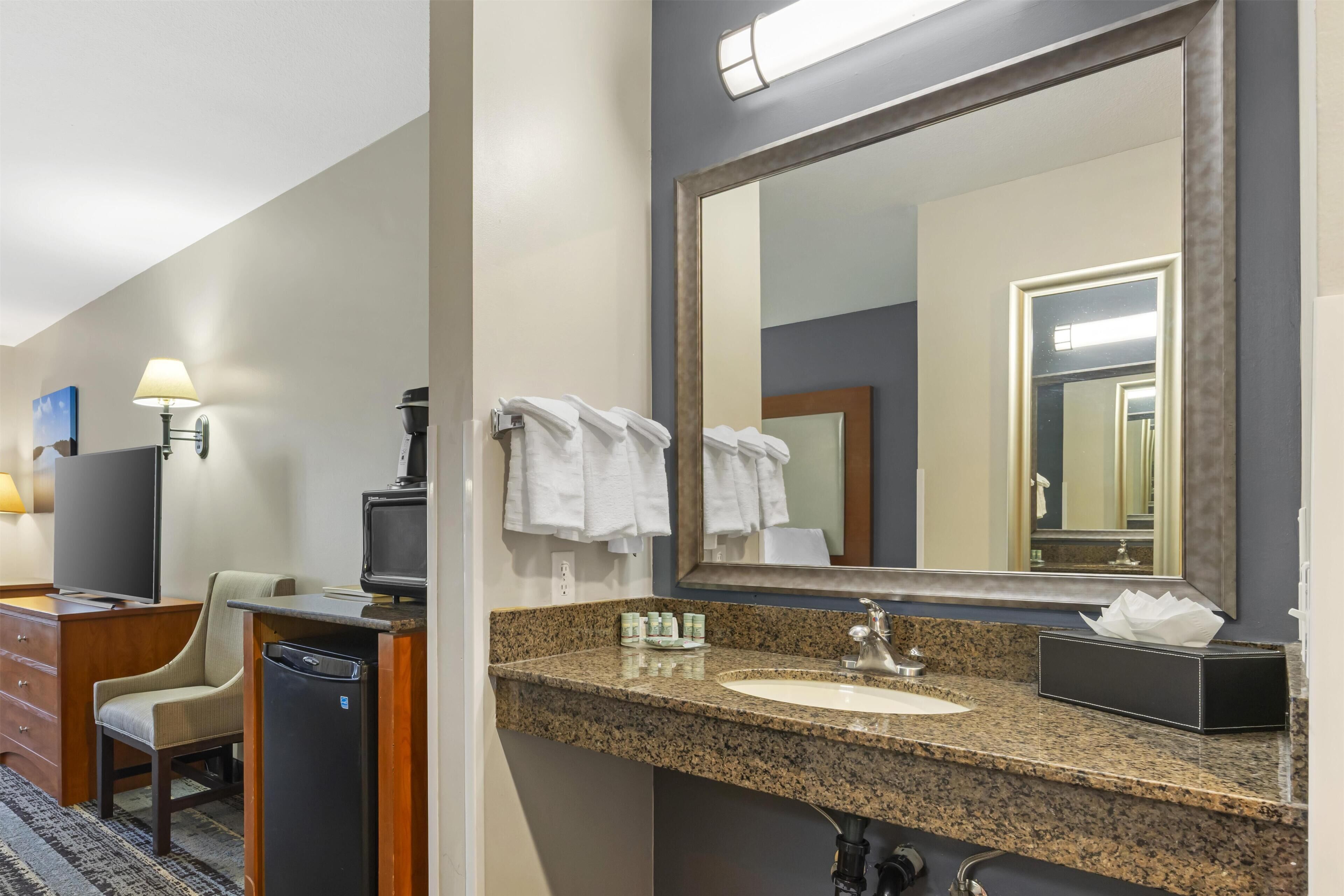 A hotel bathroom with a granite countertop sink, a large mirror, a towel rack, a chair, a television, and a coffee machine.
