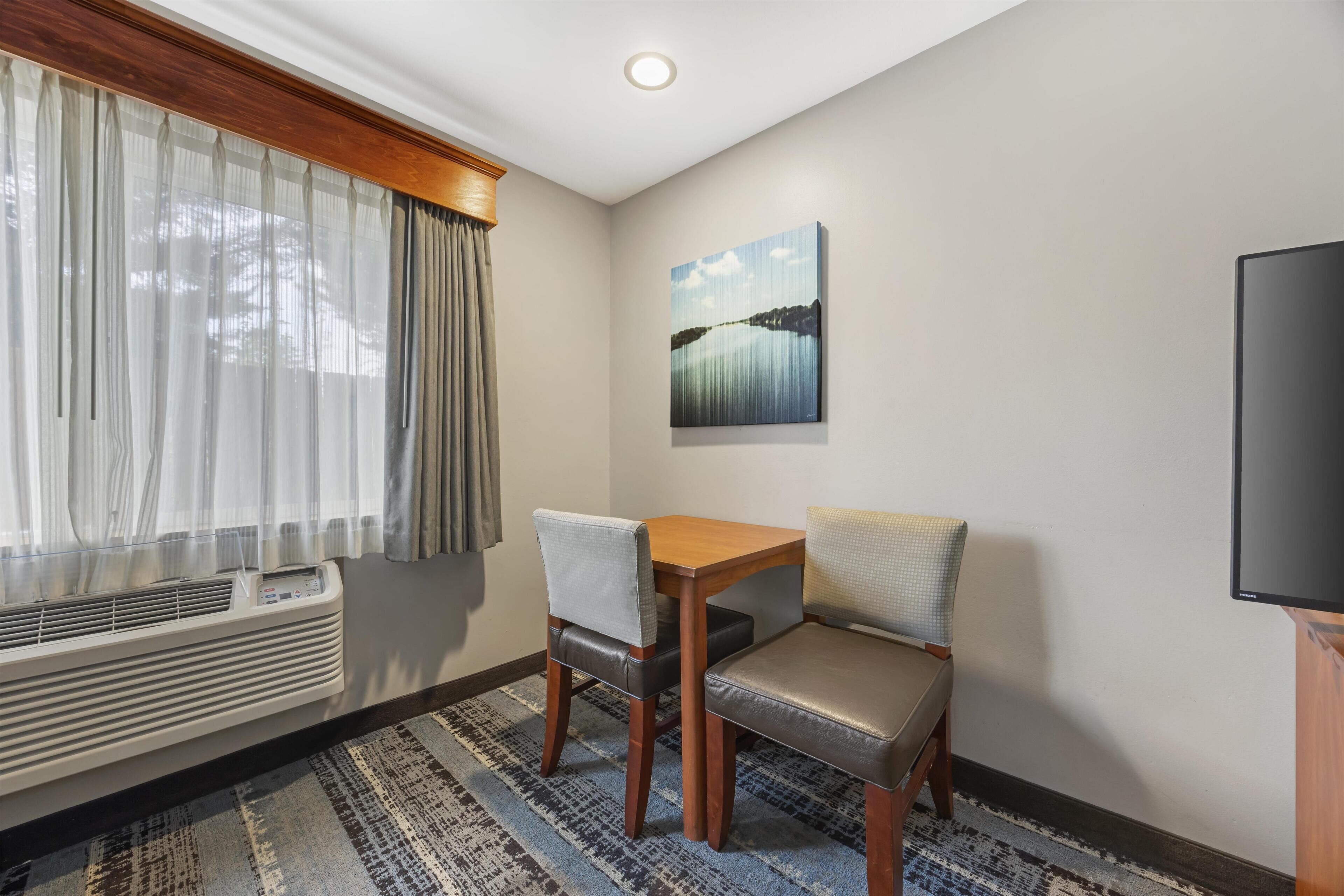 A hotel room with a dining table, two chairs, a painting on the wall, a large window with curtains, and a flat-screen TV.
