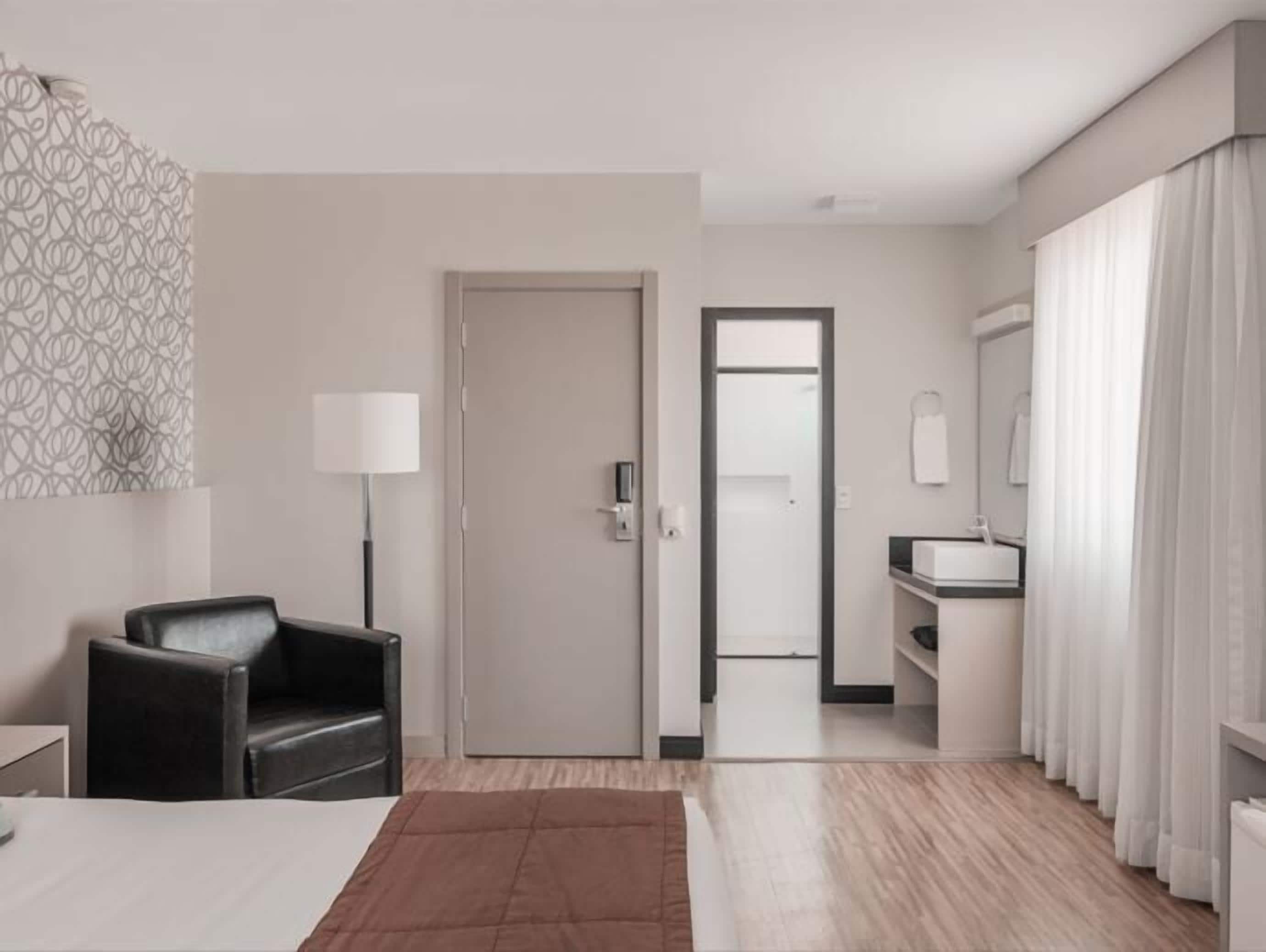 A hotel room with a bed, a desk, a chair, a lamp, a bathroom with a mirror, and a door leading to another room.