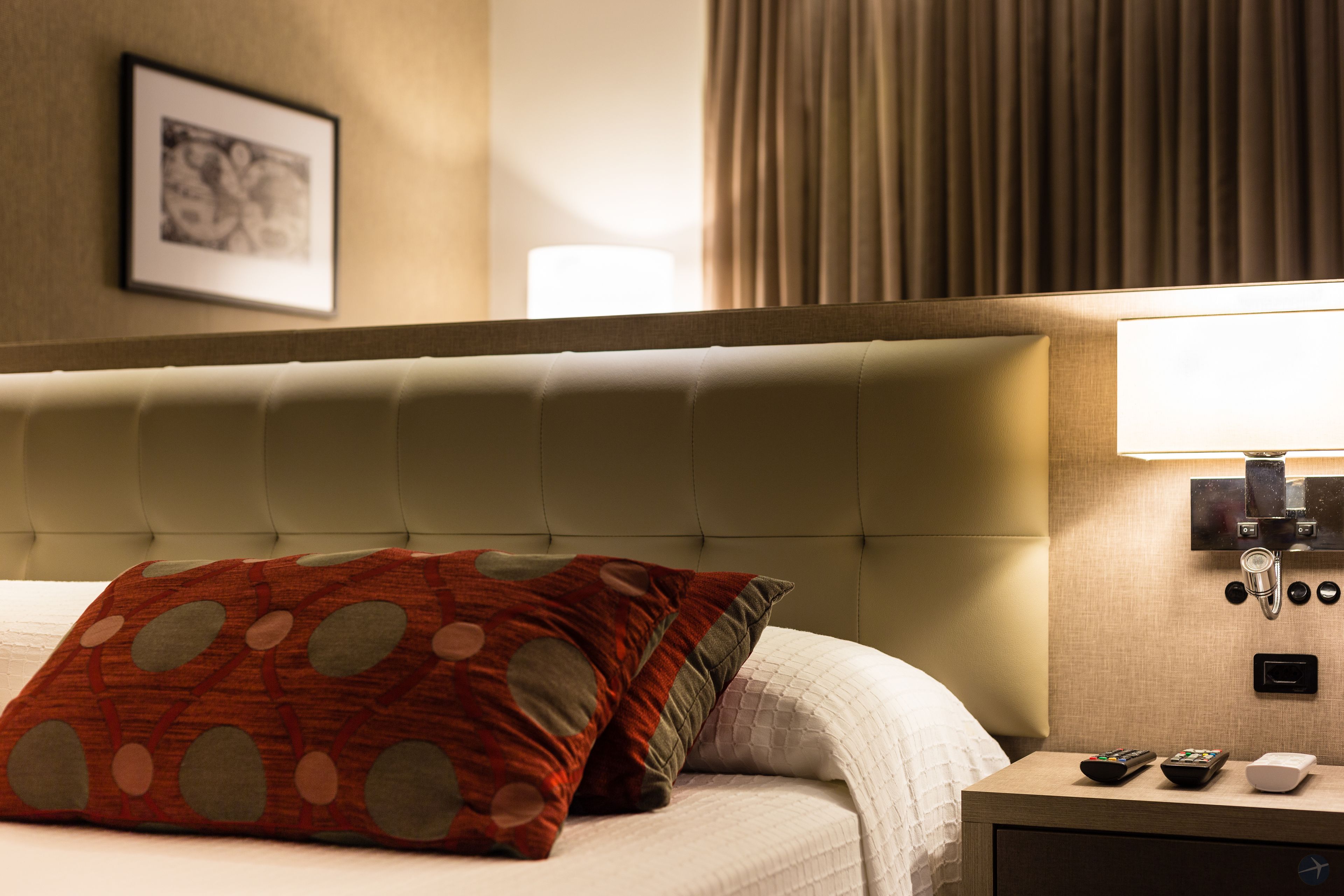 A hotel room with a tufted headboard, a bed with a white comforter, a red and grey patterned pillow, a bedside table with a lamp, and a framed picture on the wall.
