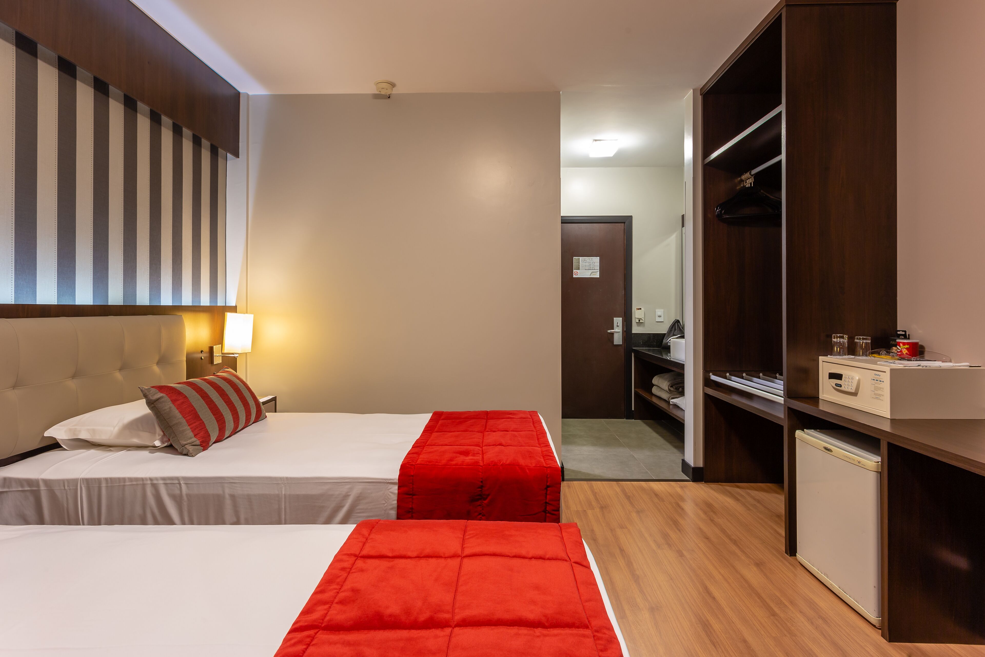 A hotel room with two beds, a wardrobe, a desk with a computer, and a small refrigerator.