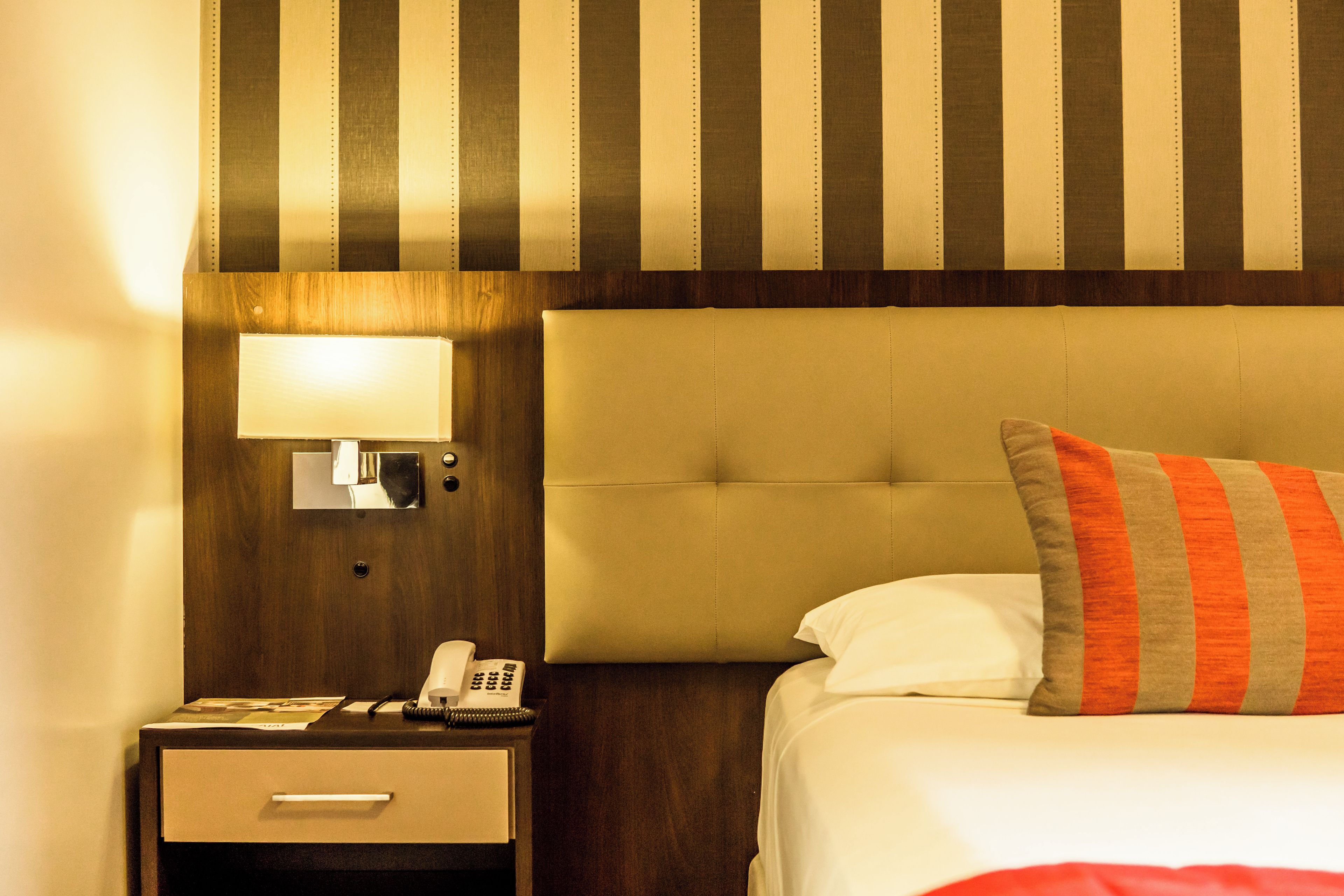 Quarto de hotel com cama, mesa de cabeceira, abajur e telefone.