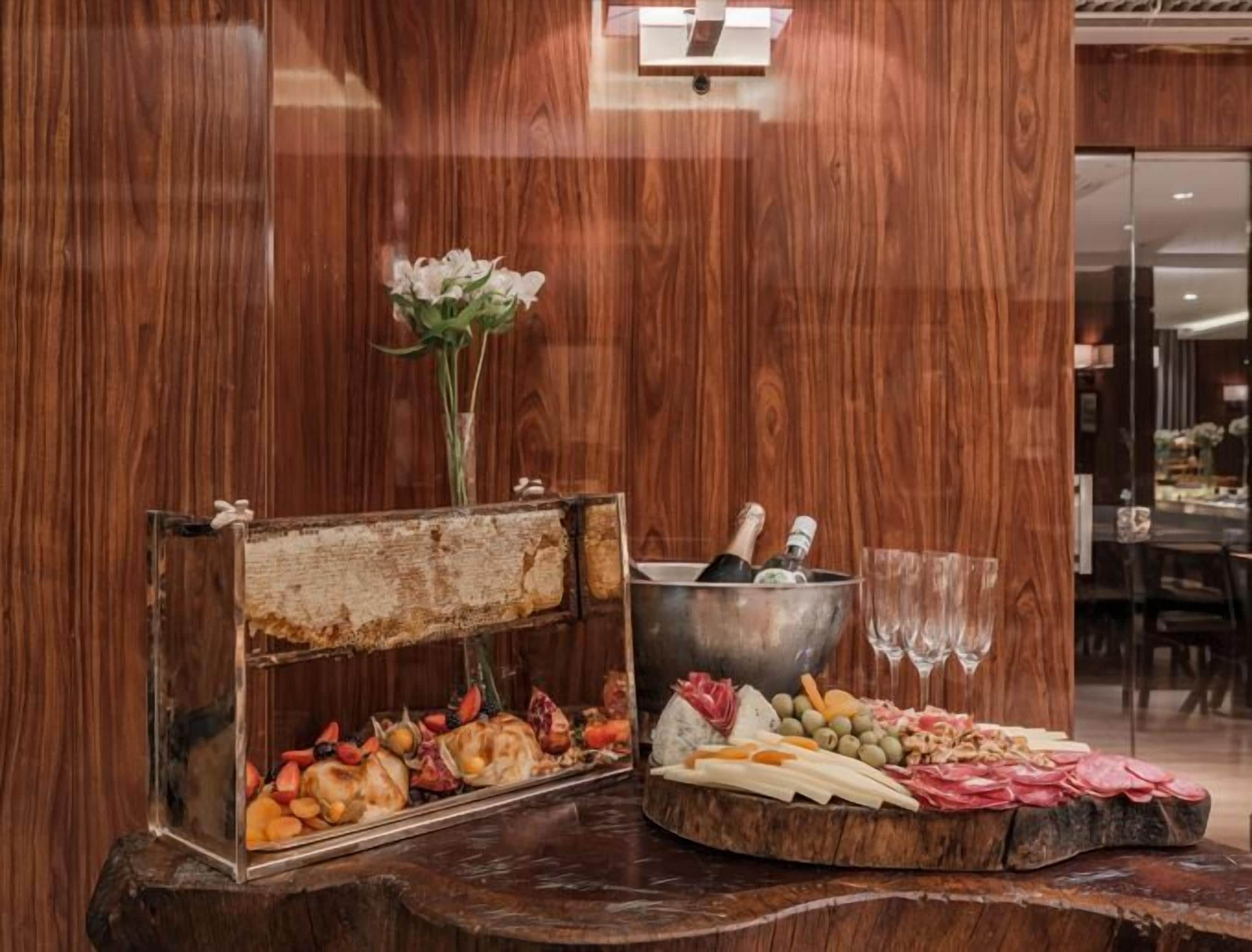 A wooden buffet table with a floral centerpiece, a chilled beverage bucket, and a variety of cheeses and meats.