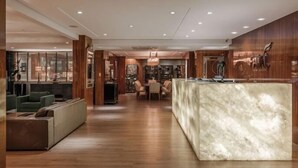 Um hall de hotel moderno com um balcão de recepção de mármore, painéis de madeira e uma área de estar com sofás e uma mesa de jantar.