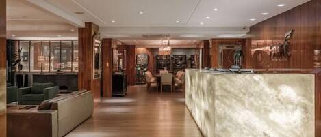 Una hall d'albergo moderna con una reception in marmo, pannelli in legno e un'area salotto con divani e un tavolo da pranzo.