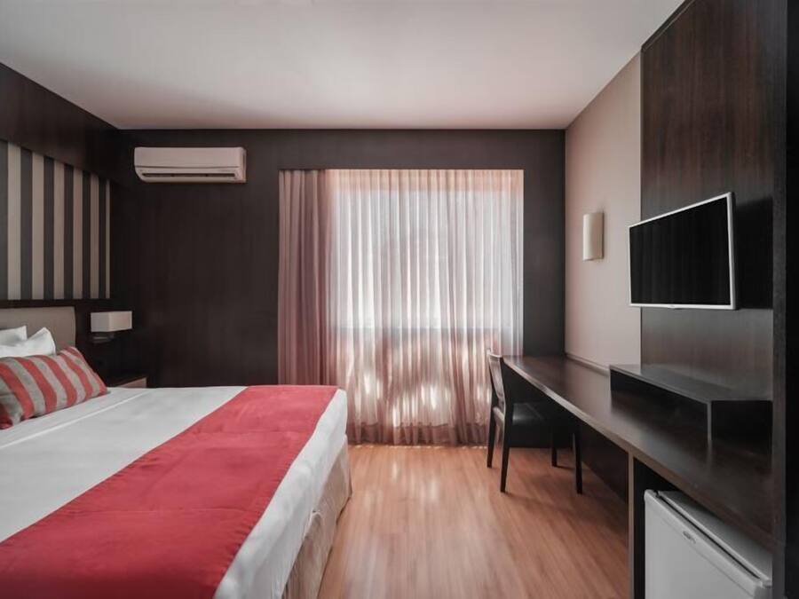 A modern hotel room with a bed, a desk, a TV, and a window with curtains.
