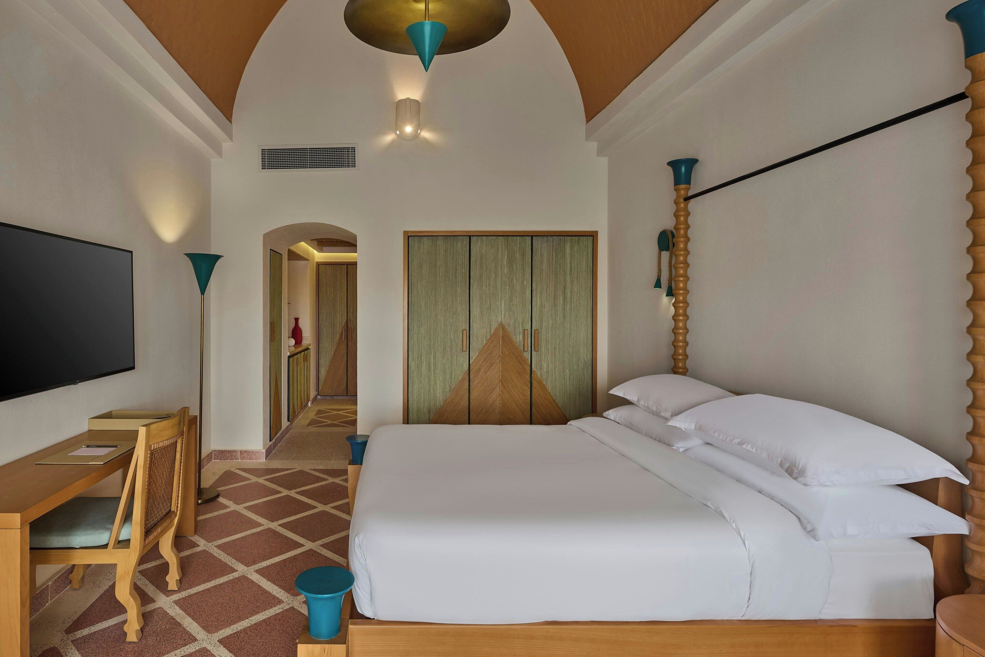 A hotel room with a bed, a desk, a chair, a TV, and a door leading to another room.