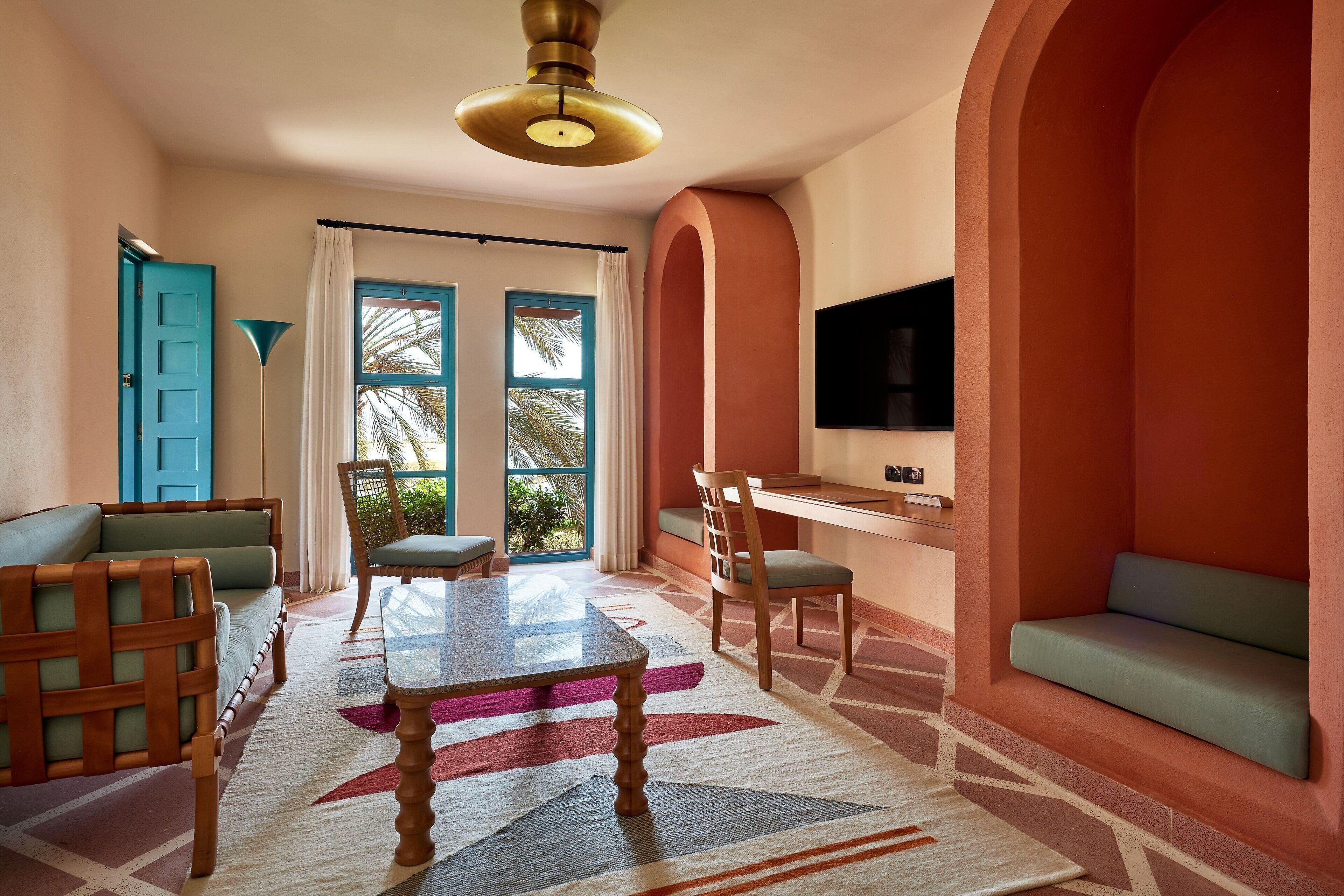 Una habitación con una mesa de madera, sillas, un sofá, un televisor montado en la pared y un ventanal con cortinas.