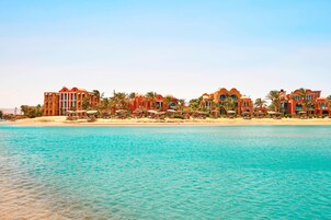 Un hotel frente al mar con varias construcciones, palmeras y una playa arenosa.