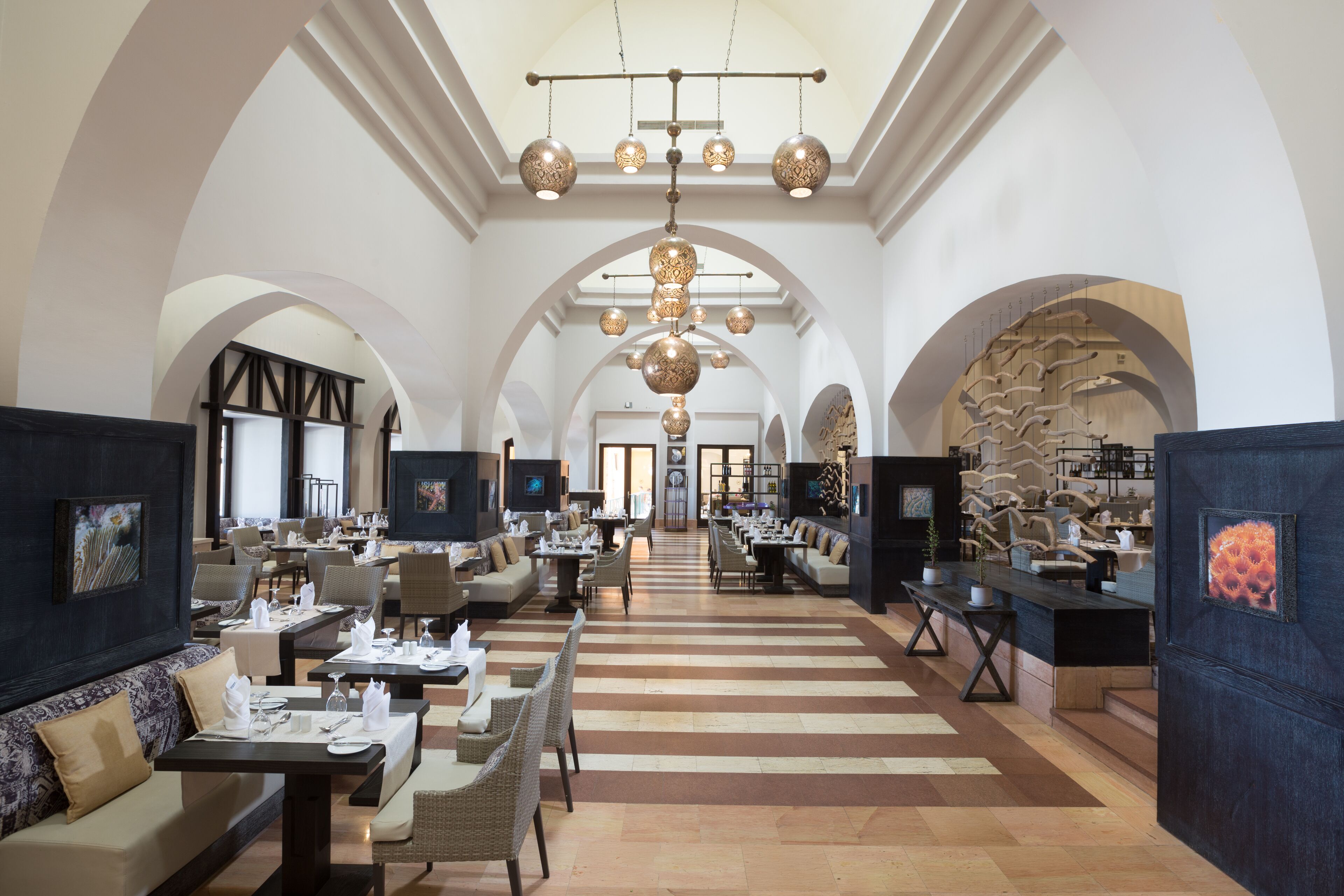 A spacious hotel lobby with high ceilings, arched doorways, and a central chandelier. Tables and chairs are arranged for dining, and artwork is displayed on the walls.