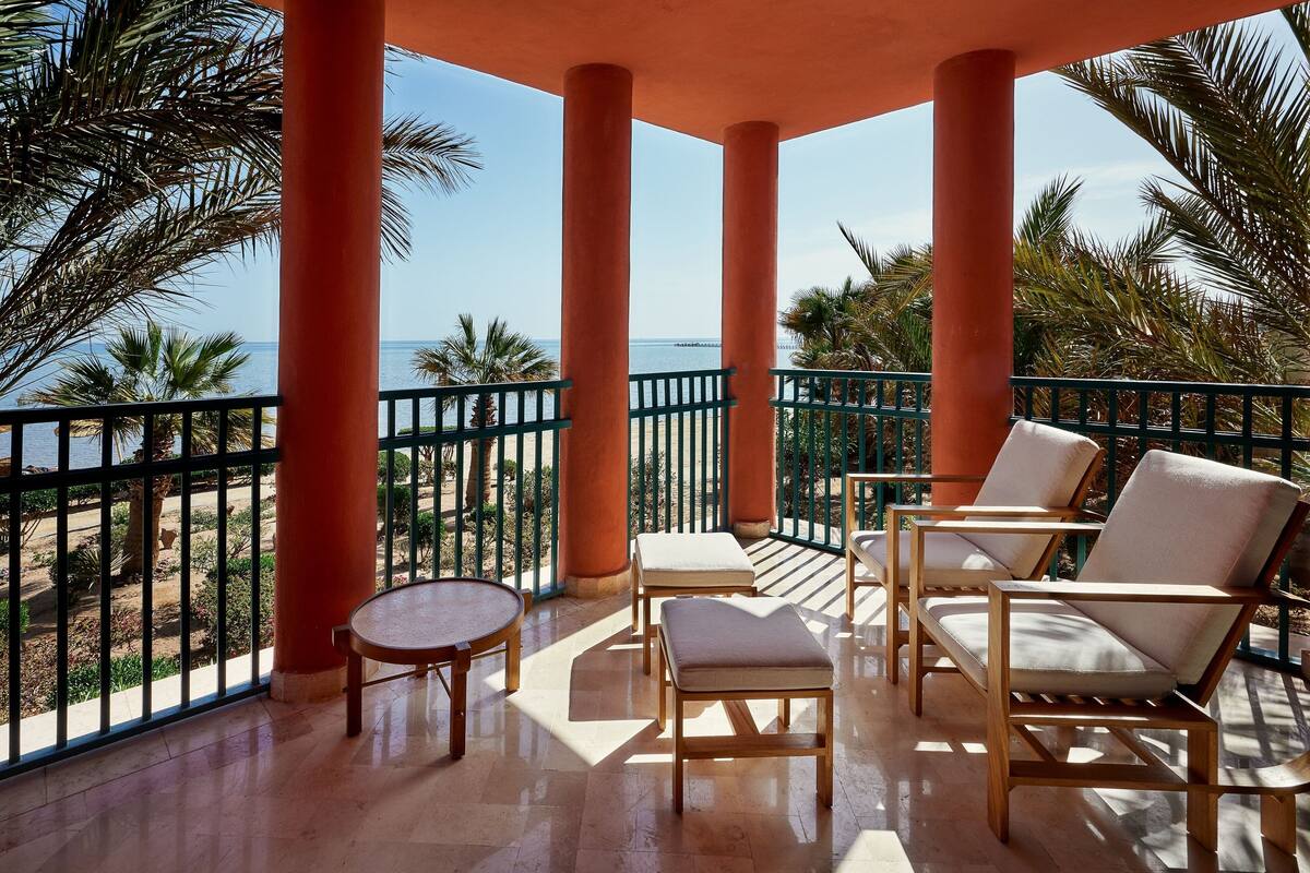 A balcony with wooden chairs and a small table, overlooking a beach and palm trees.