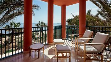 A balcony with wooden chairs and a small table, overlooking a beach and palm trees.