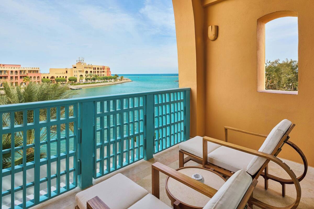 A balcony with a view of the sea and buildings, featuring a wooden table and two lounge chairs.