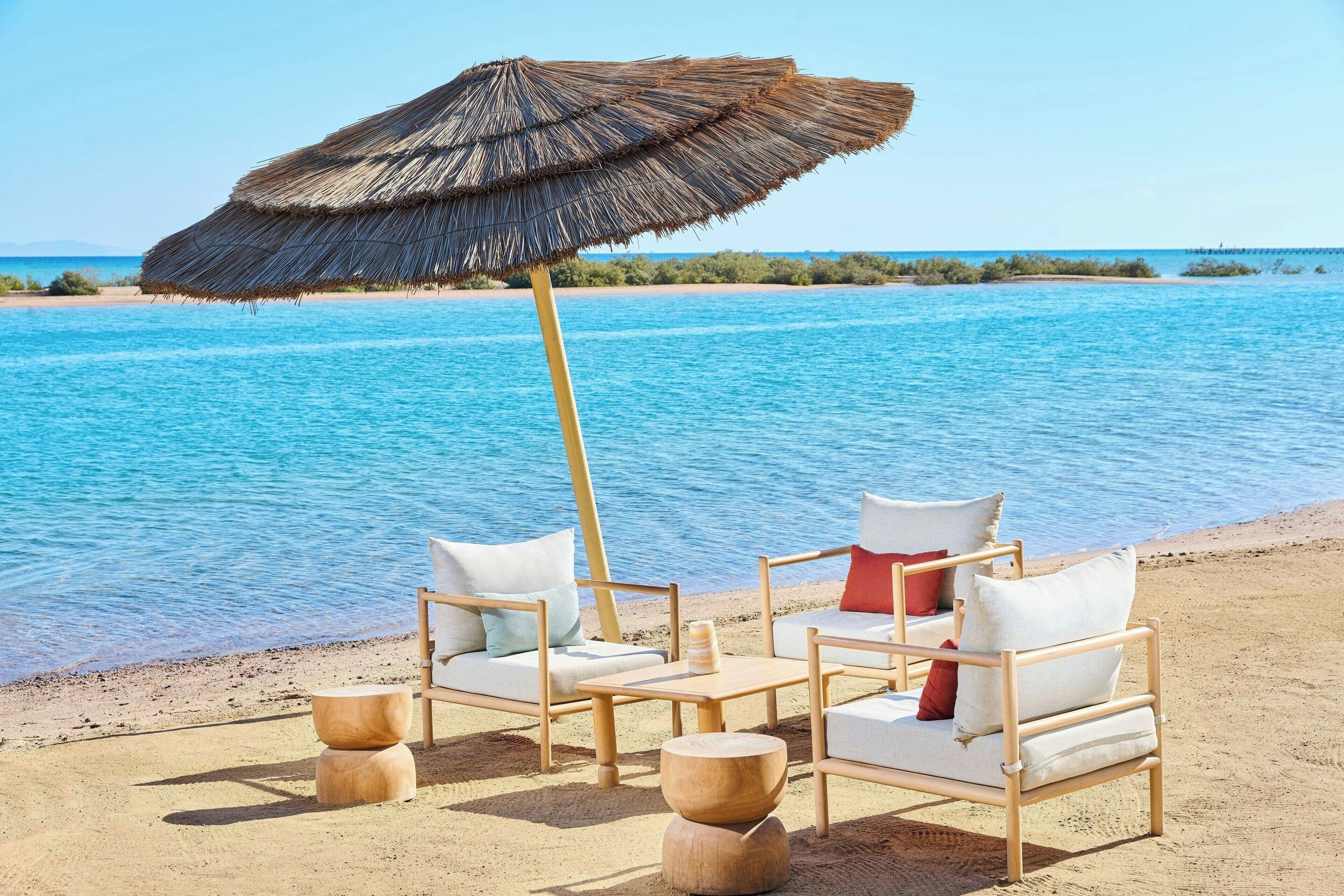 Un paisaje de playa con dos sillas de madera, una sombrilla de paja, una mesa de madera y un taburete pequeño de madera.