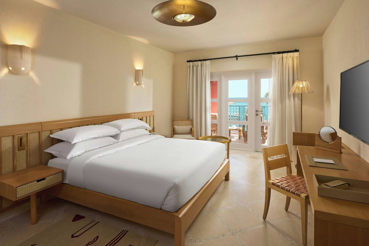 A hotel room with a large bed, a desk with a chair, a TV, and a balcony with curtains.