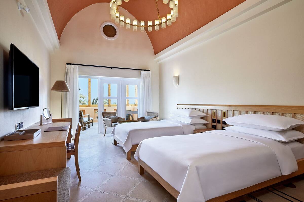 A hotel room with two beds, a desk, a TV, and a large window with curtains.